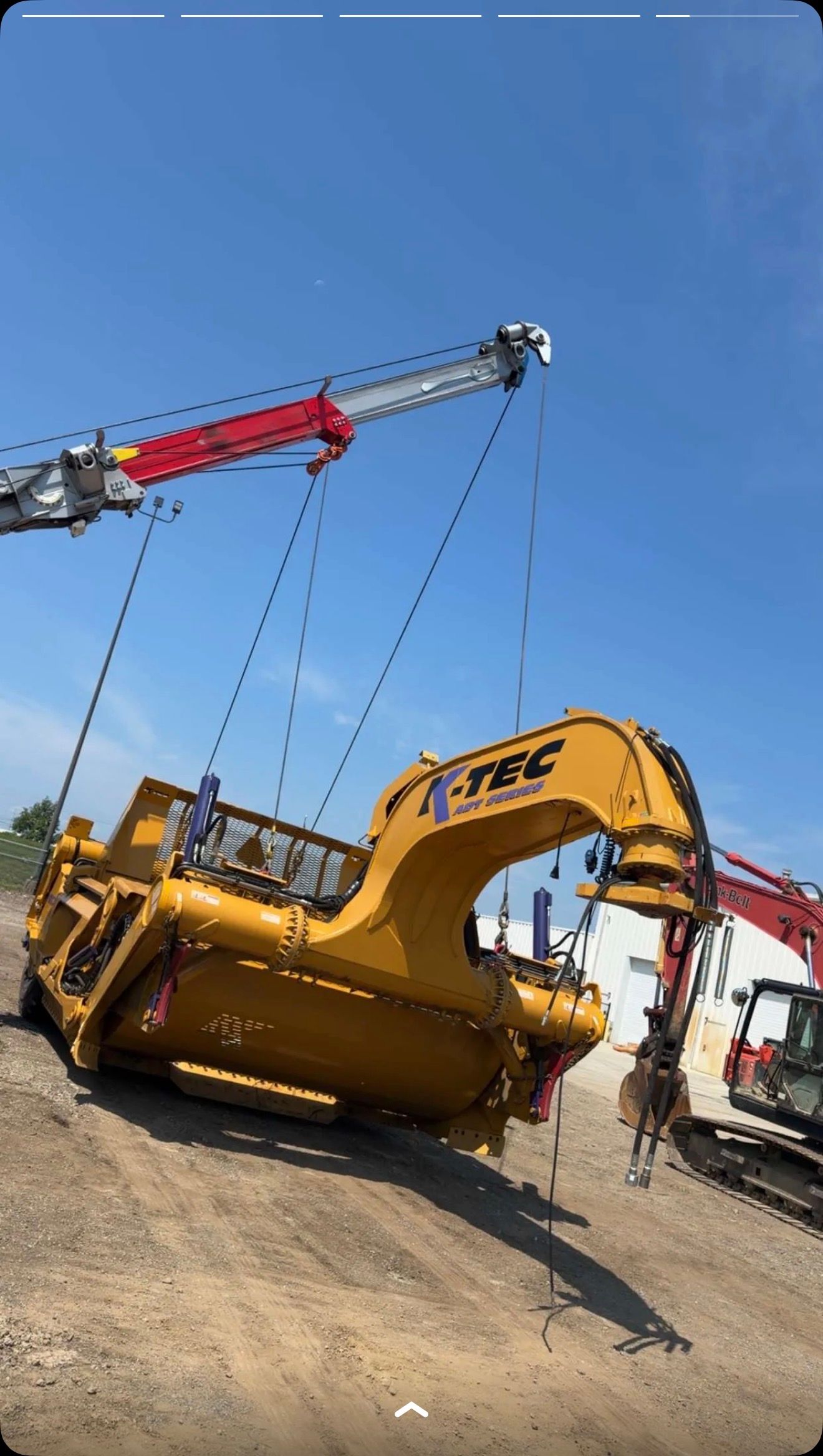 Yellow excavator being lifted by a crane at a construction site under a clear blue sky