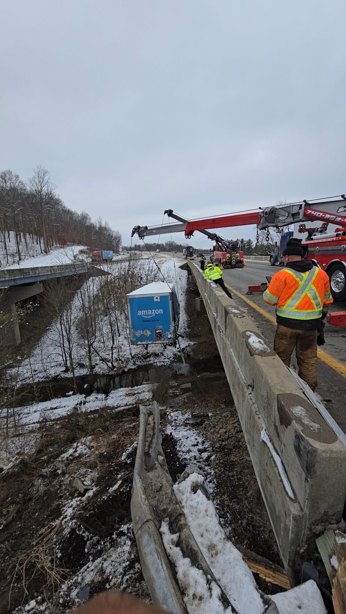 Workers in orange vests on a snowy roadside beside a blue portable toilet and damaged guardrail
