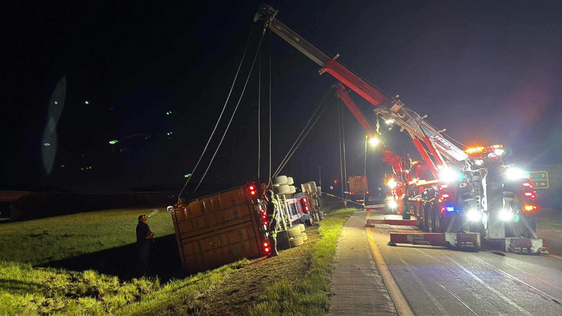 Tow truck lifting a tipped trailer on a road at night, with emergency lights flashing.