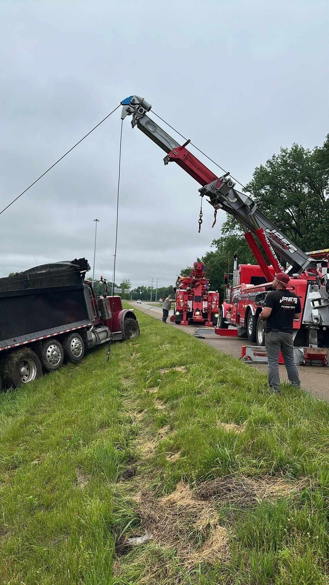 Tow truck hoisting a semi-trailer from a roadside ditch beside an overturned red truck.