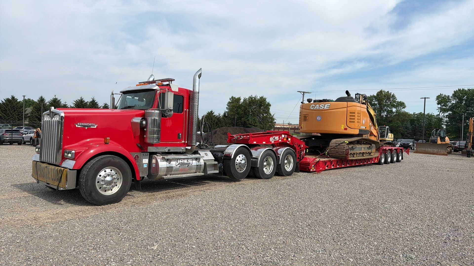 Red semi truck hauling a large orange construction machine on a flatbed trailer in a gravel lot.