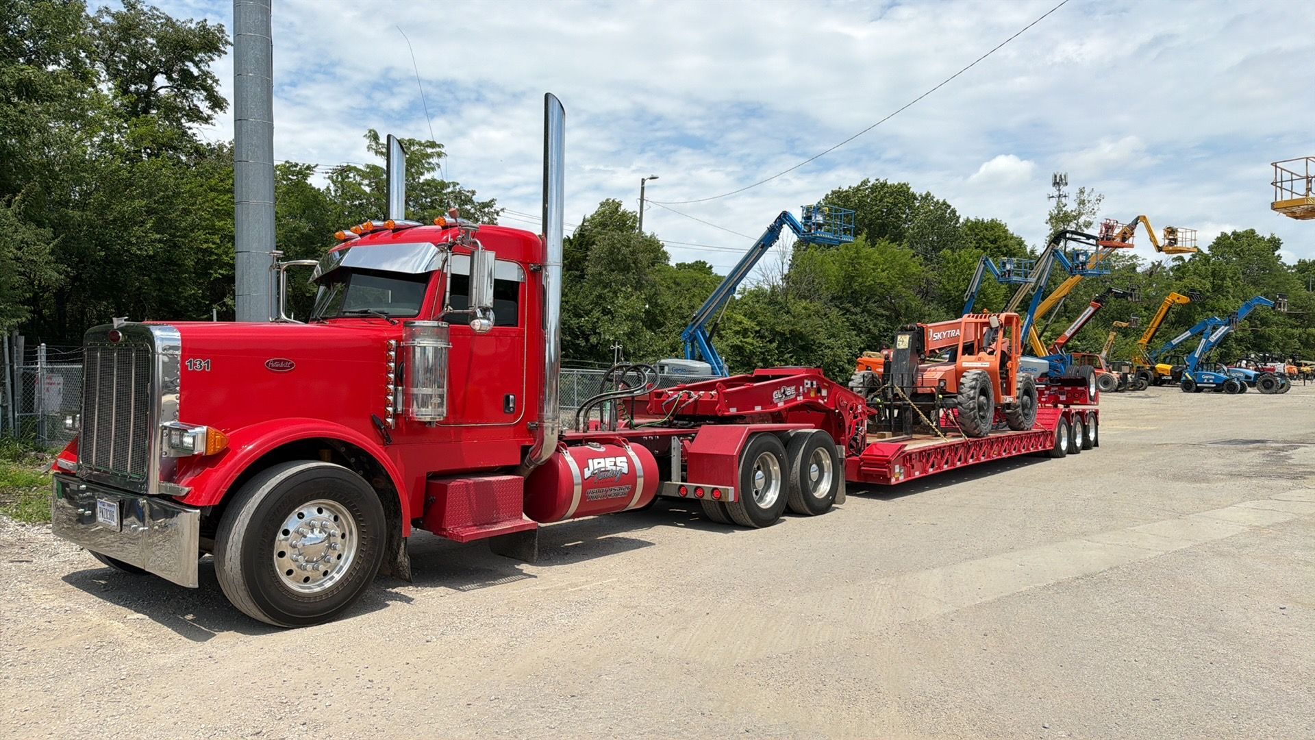 Red semi truck hauling an orange excavator on a flatbed trailer beside a road