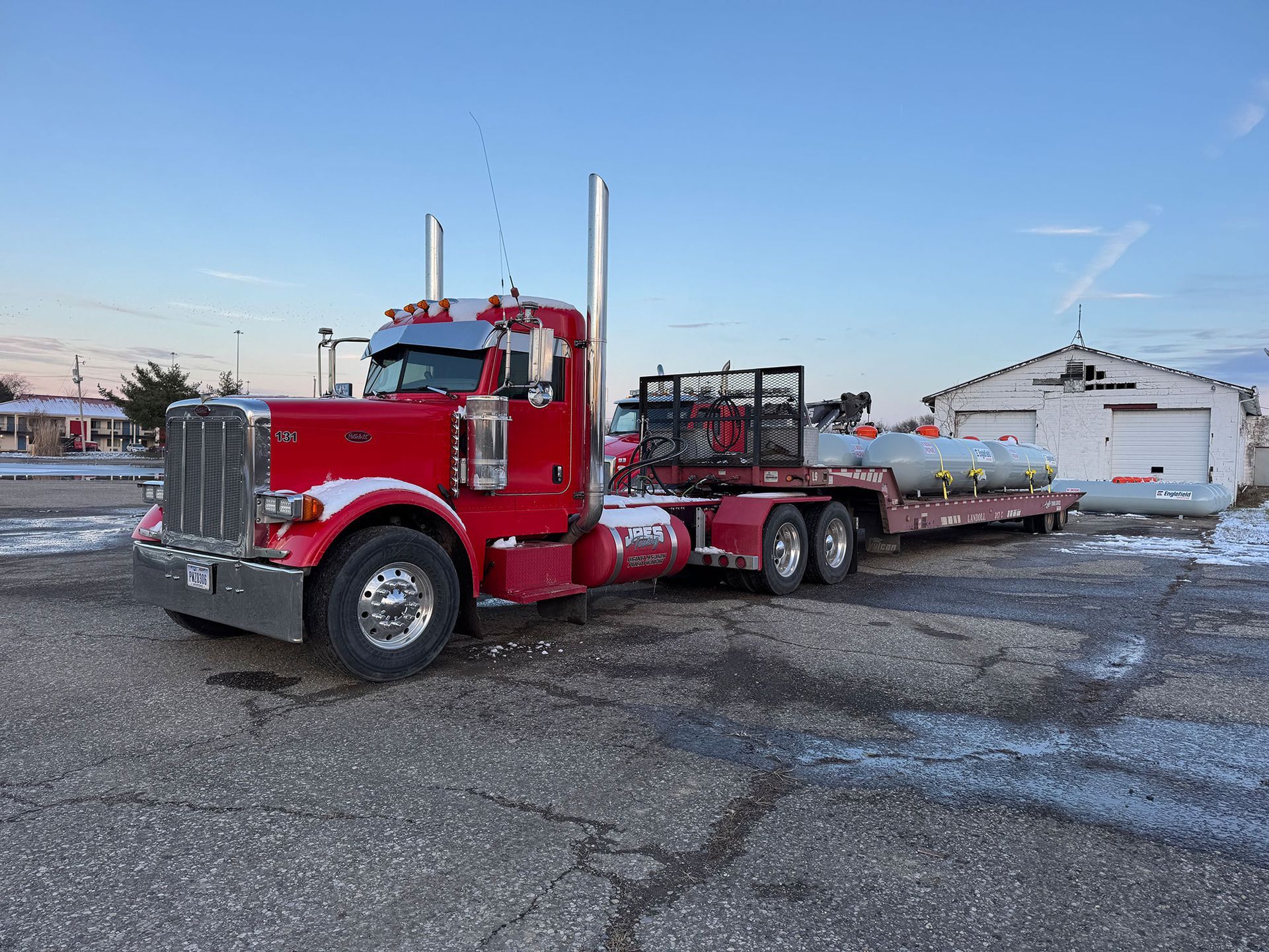 Red semi truck hauling a flatbed trailer on a snowy lot near buildings at dusk