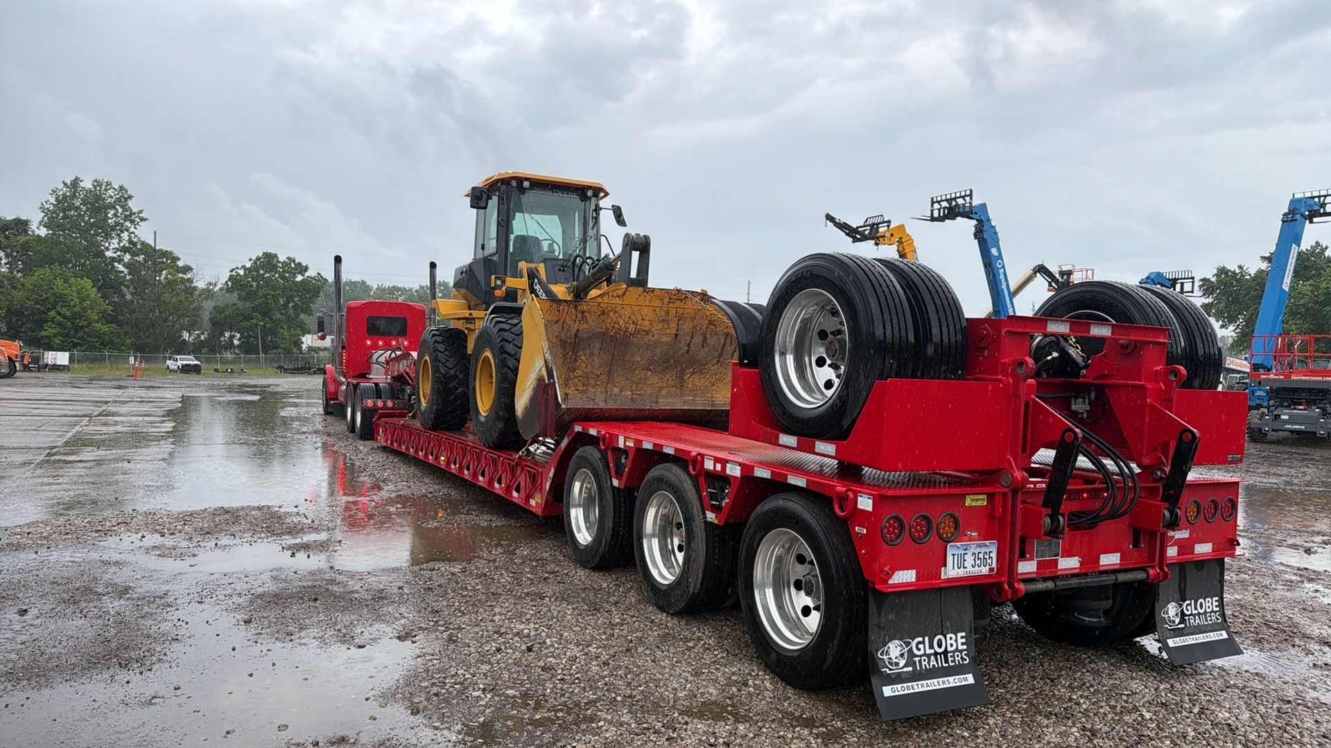 Red flatbed trailer hauling a yellow loader and spare tire through a wet parking lot.