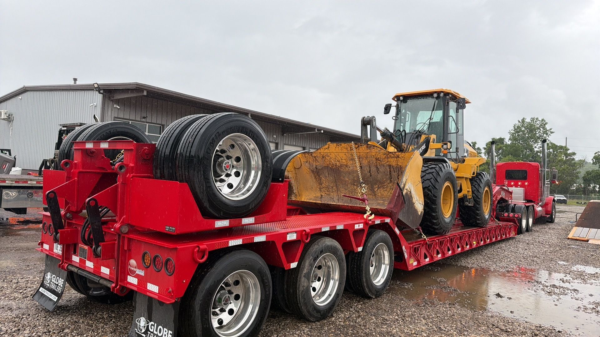 Red flatbed trailer hauling a yellow excavator in a gravel yard, with a red truck in the background