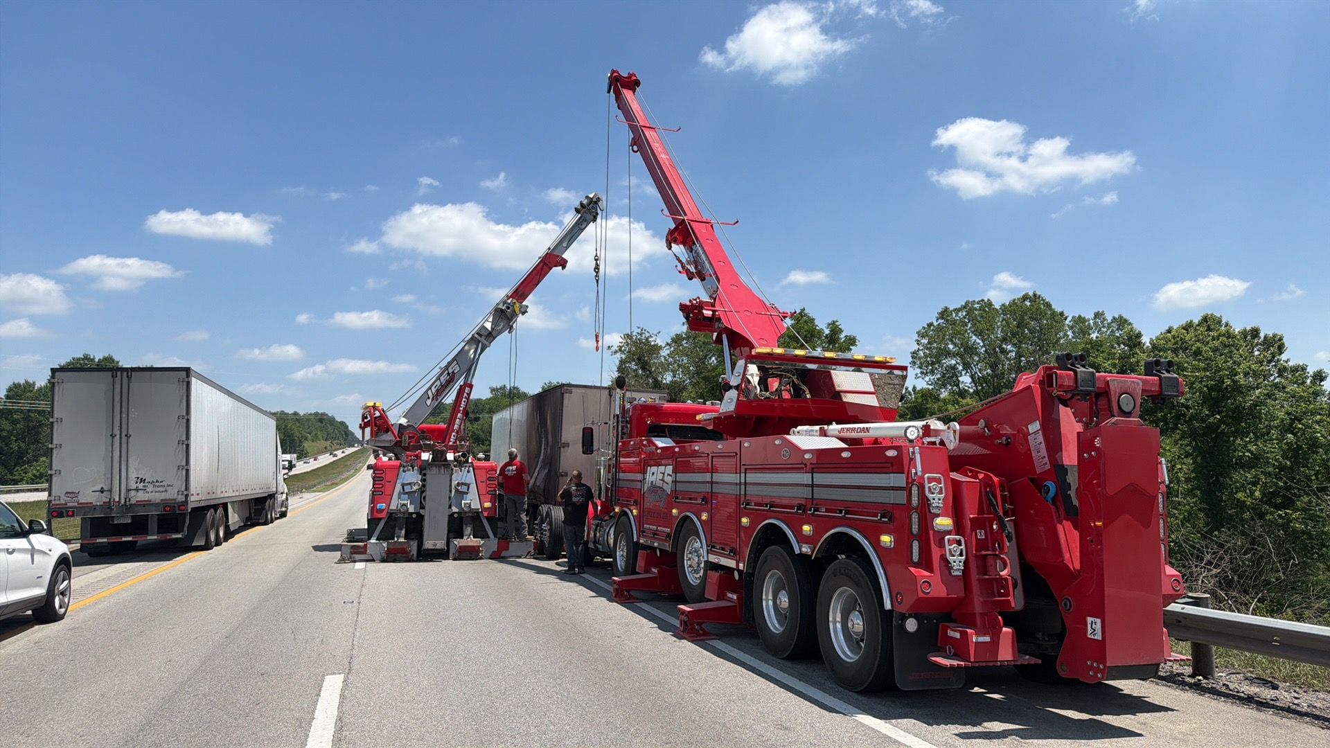 Red tow truck lifting a semi-trailer on a highway shoulder under a blue sky