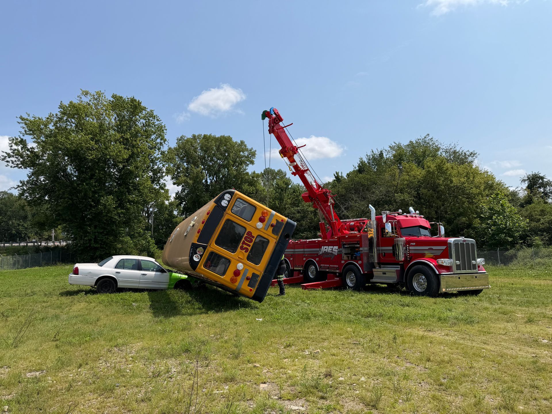 Tow truck lifting a yellow school bus on a grassy field beside a white car.