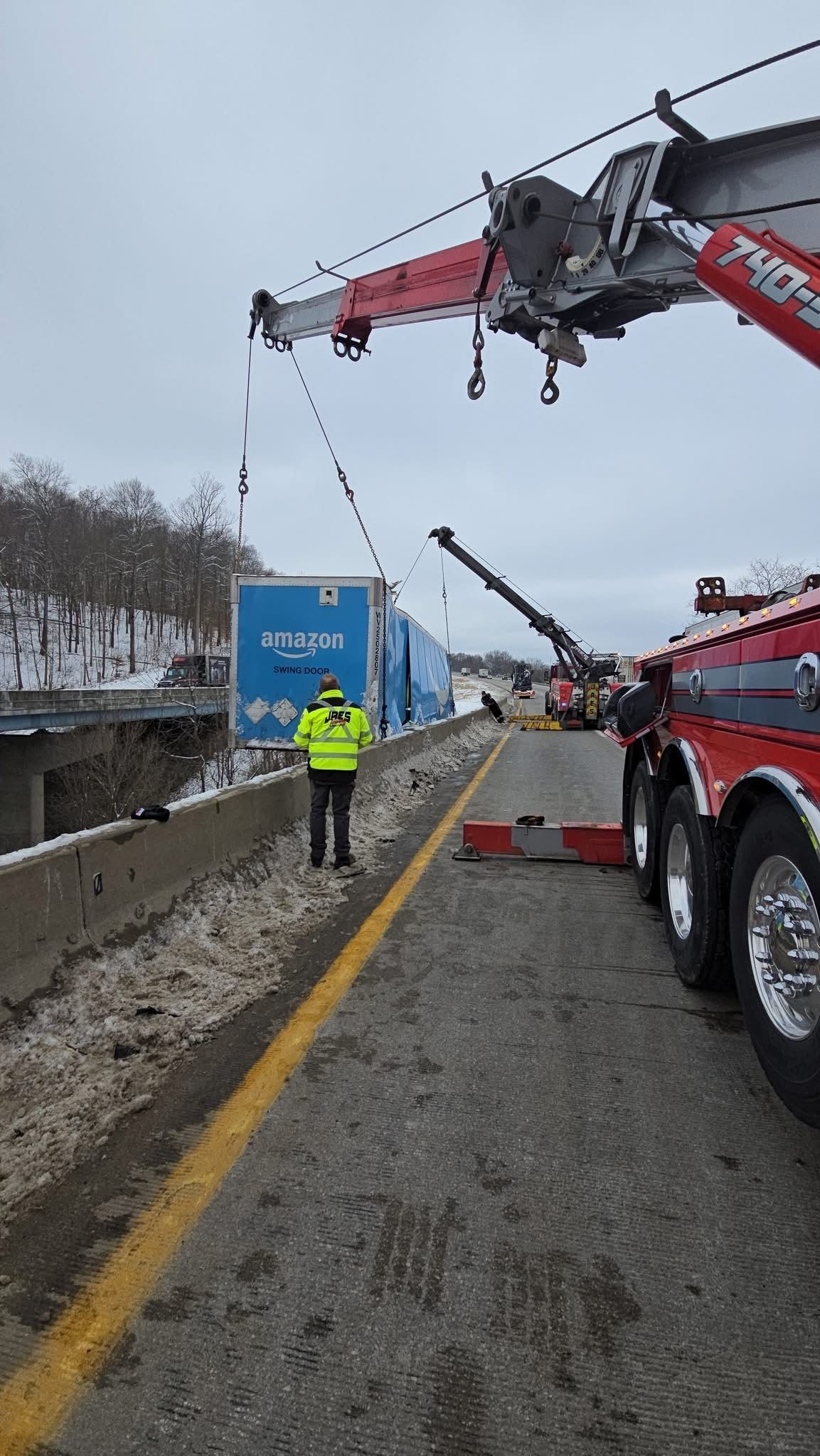 Tow truck lifting a blue vehicle from a snowy roadside, with a worker nearby and a firefighter truck overhead