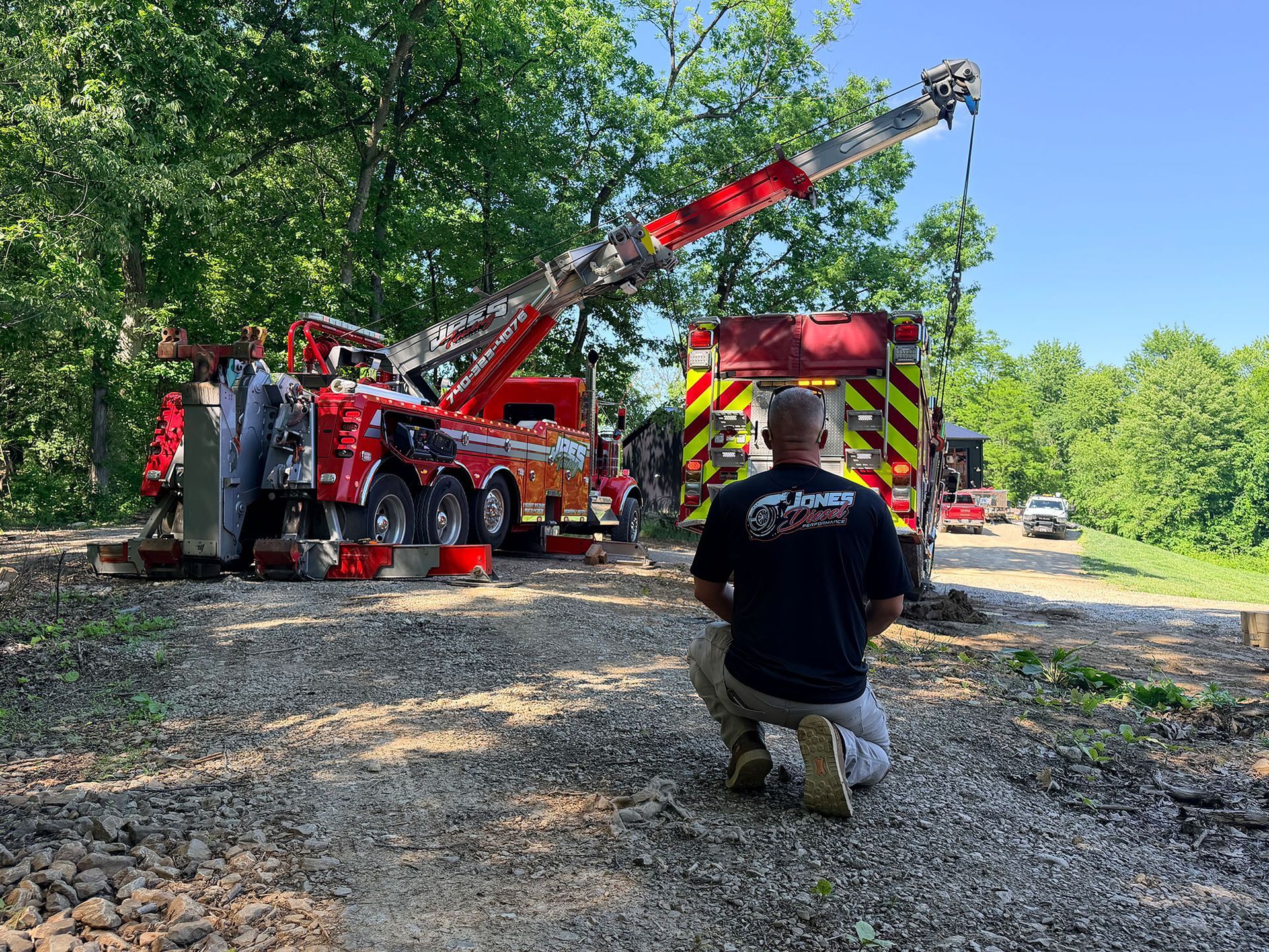 Fire truck and extended ladder on a wooded roadside, with a kneeling firefighter in the foreground.