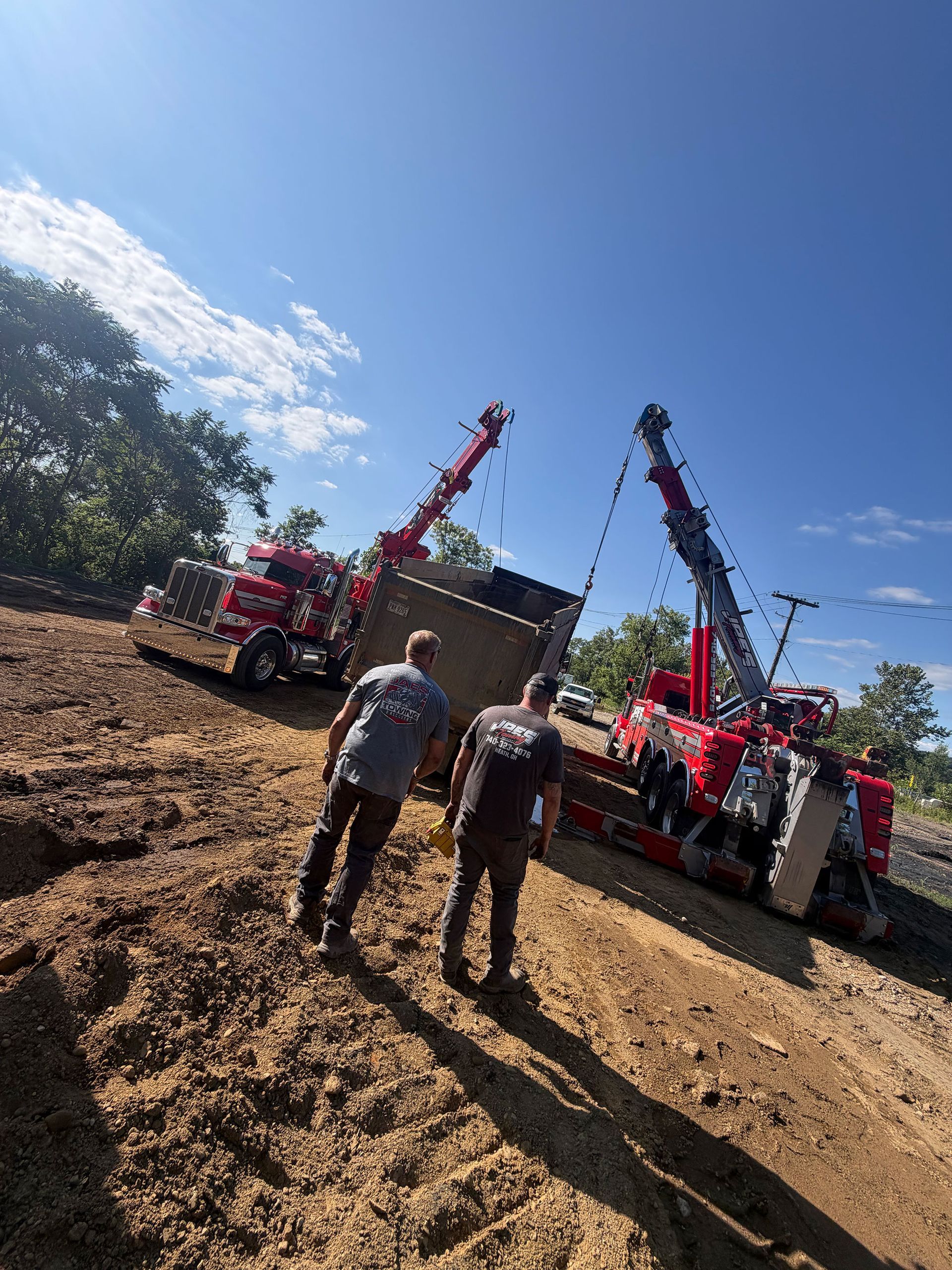 Two workers stand between two red crane trucks on a muddy worksite under a bright blue sky.