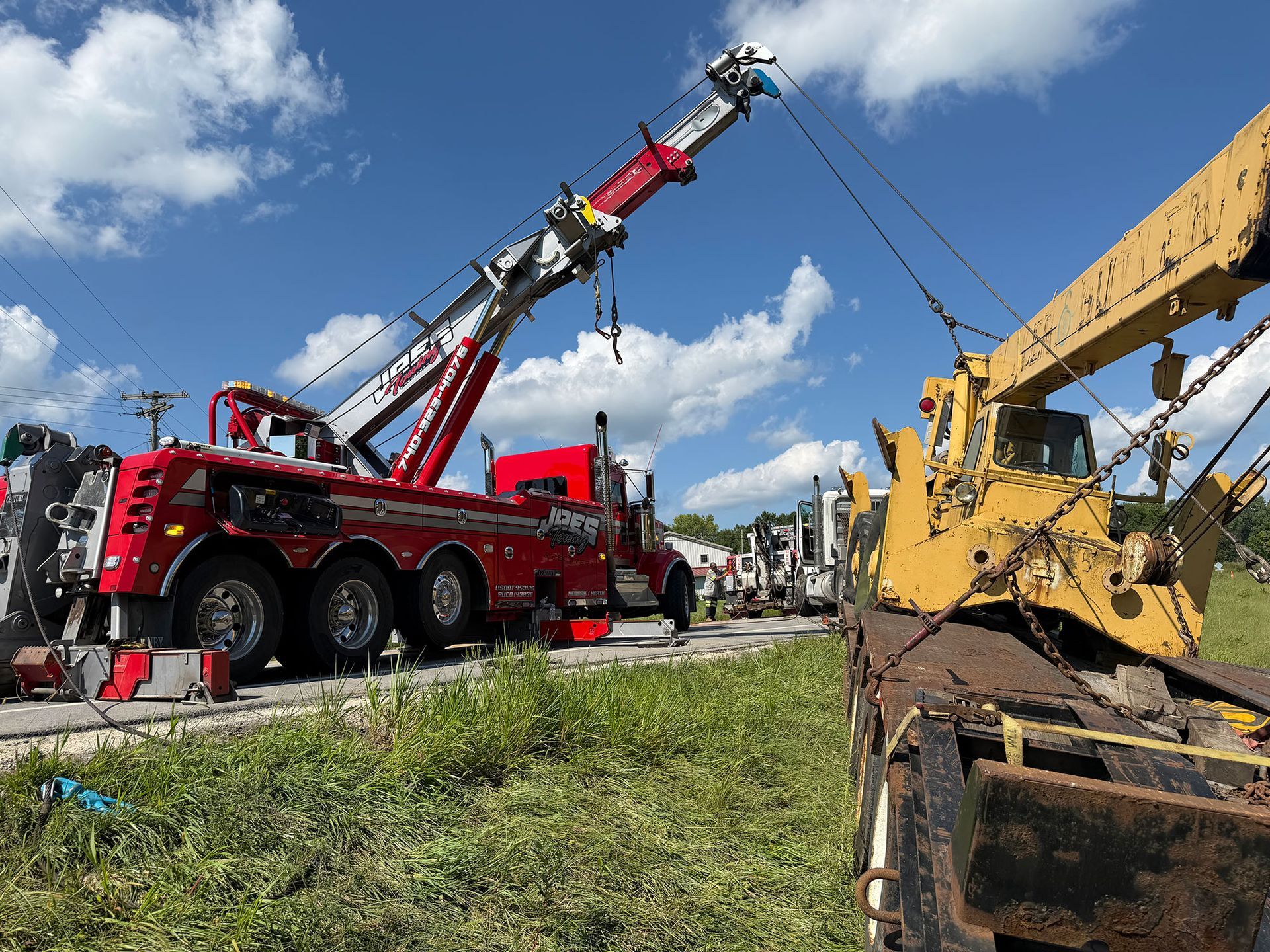 Red tow truck lifting heavy equipment beside a yellow excavator on a grassy roadside under a blue sky