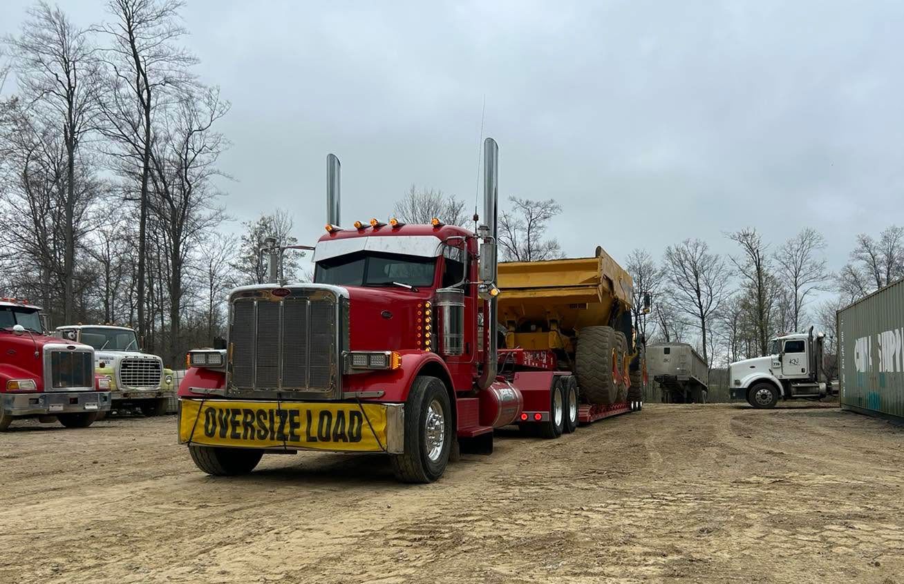 Red semi-truck with yellow dump trailer parked on muddy lot near other trucks and leafless trees