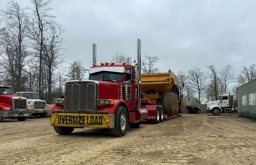Red semi-truck with a yellow dump trailer parked in a muddy lot, surrounded by other trucks and bare trees.