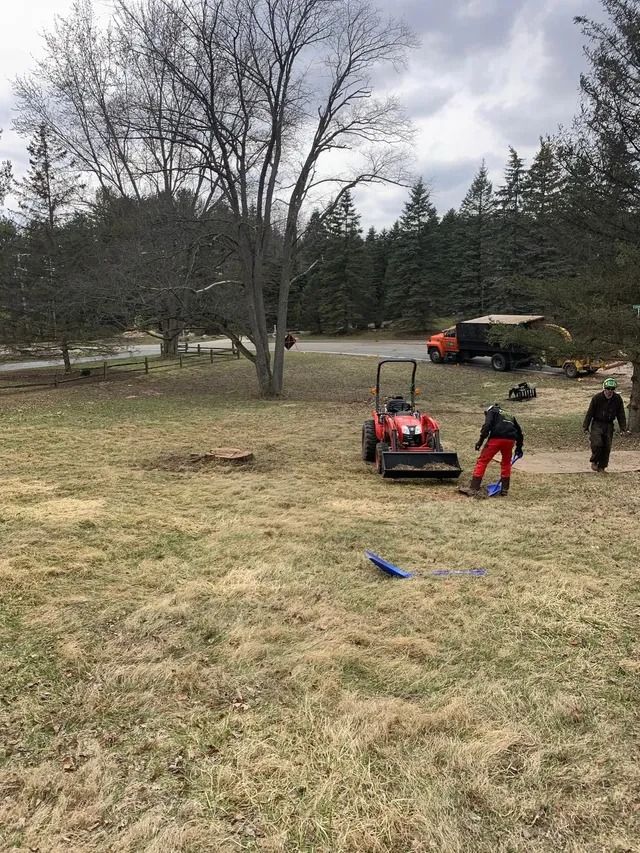Man operating a red mower, another man walking, next to truck, bare trees and brown grass.