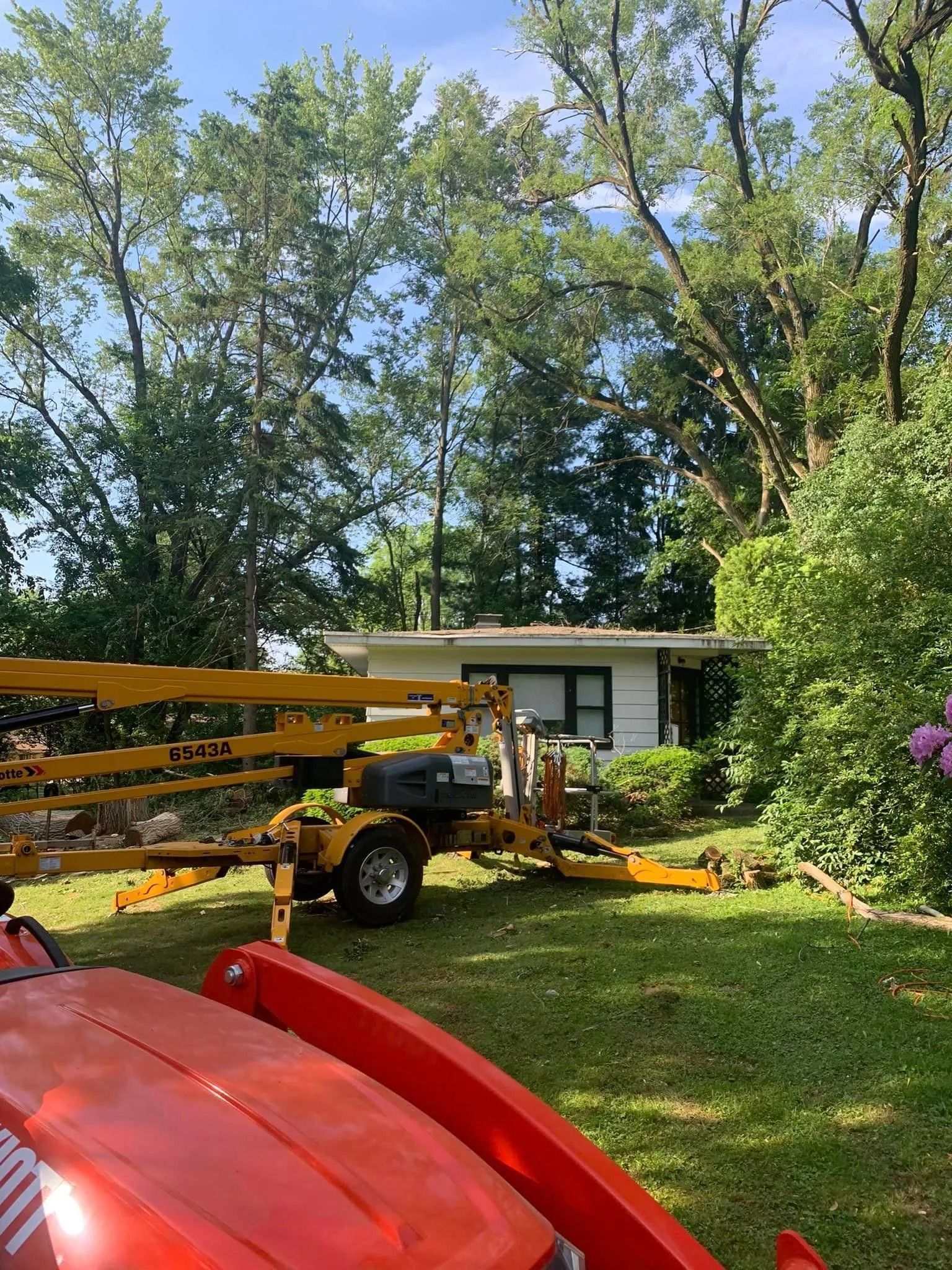 A trailer loaded with yellow equipment is in a yard with a small white building and trees in the background.