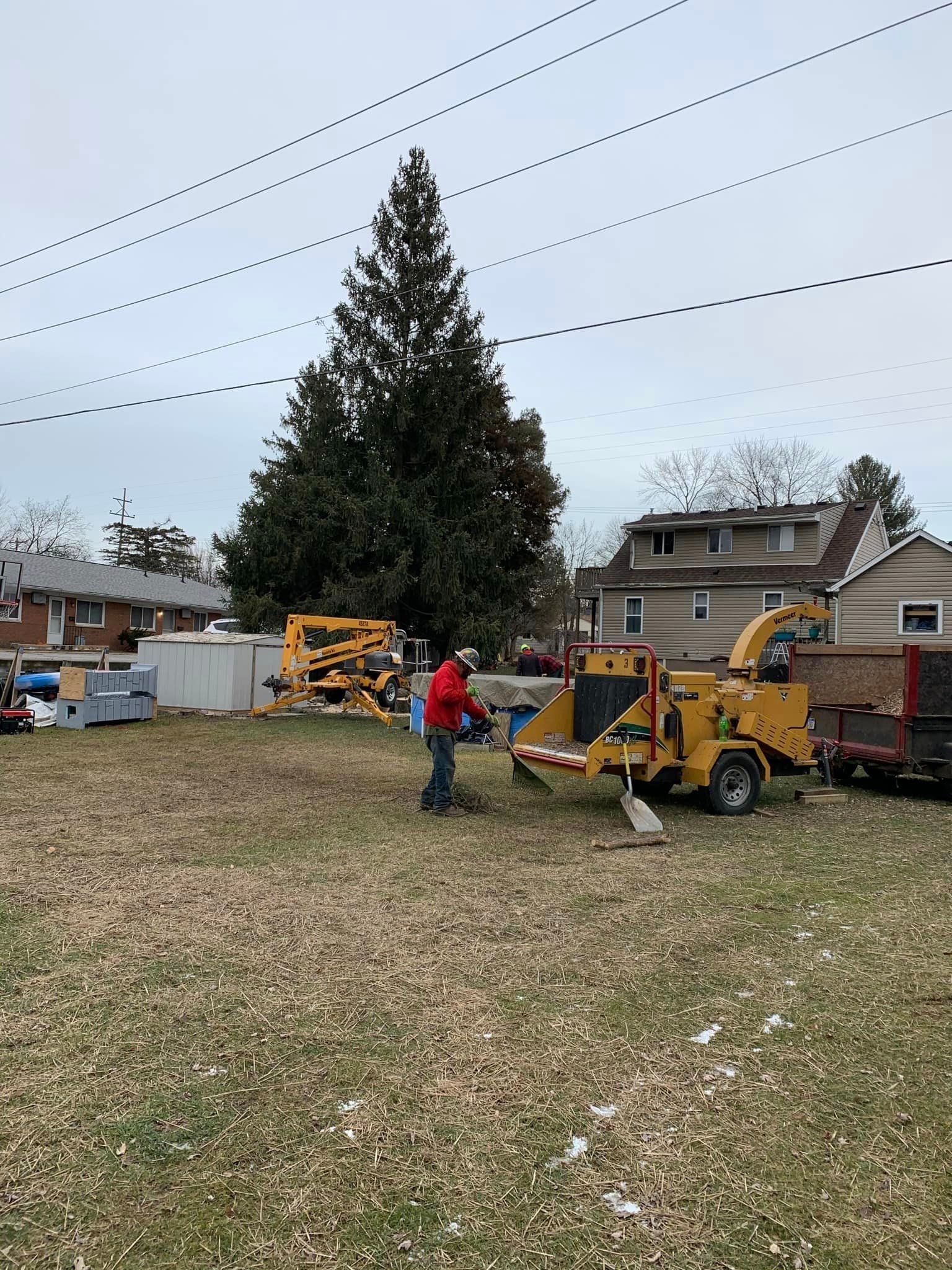 A person operating a wood chipper in a grassy yard, with a large tree and houses in the background.