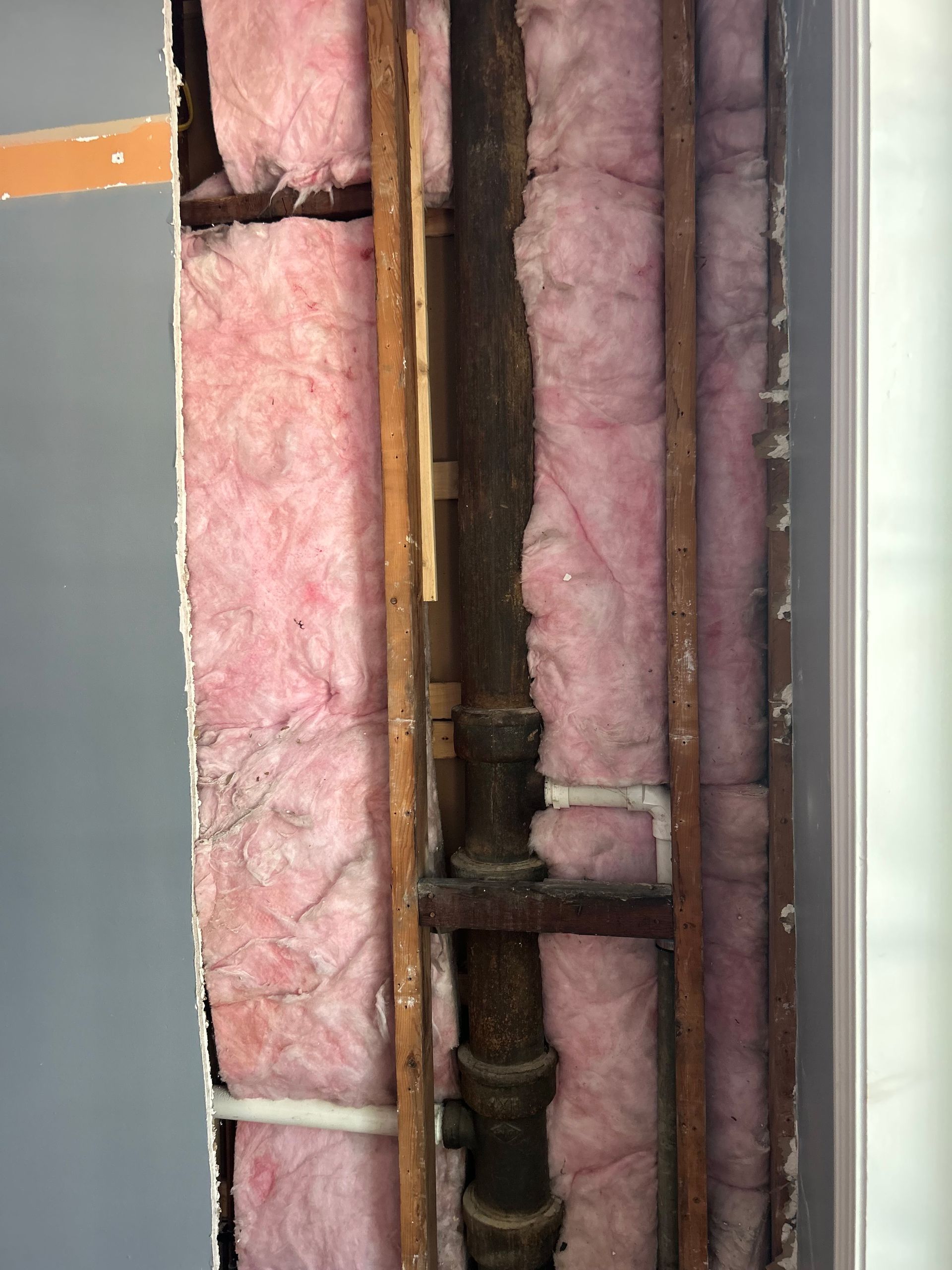 Exposed wall cavity with pink insulation, brown pipes, and wooden framing.