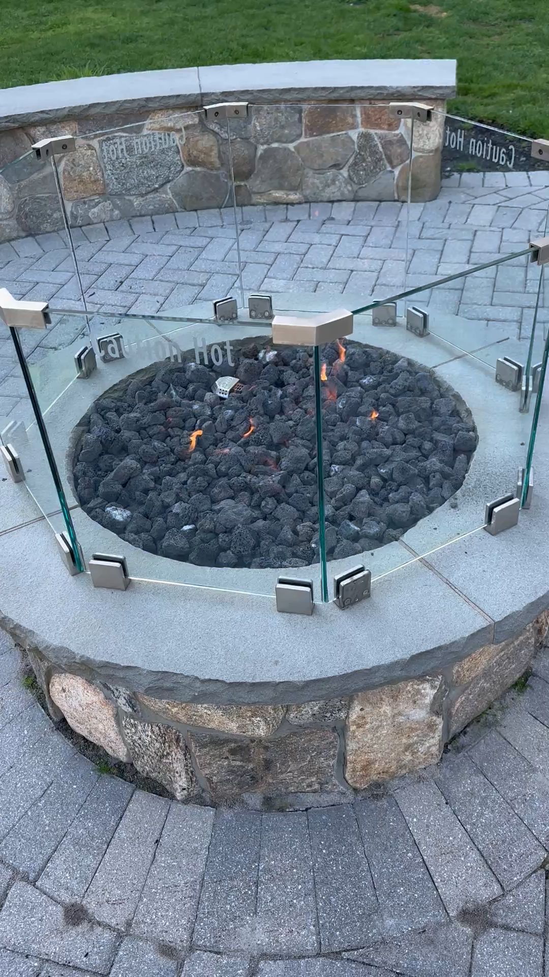 Circular fire pit with glass wind guard, surrounded by stone patio. Flames visible inside pit.