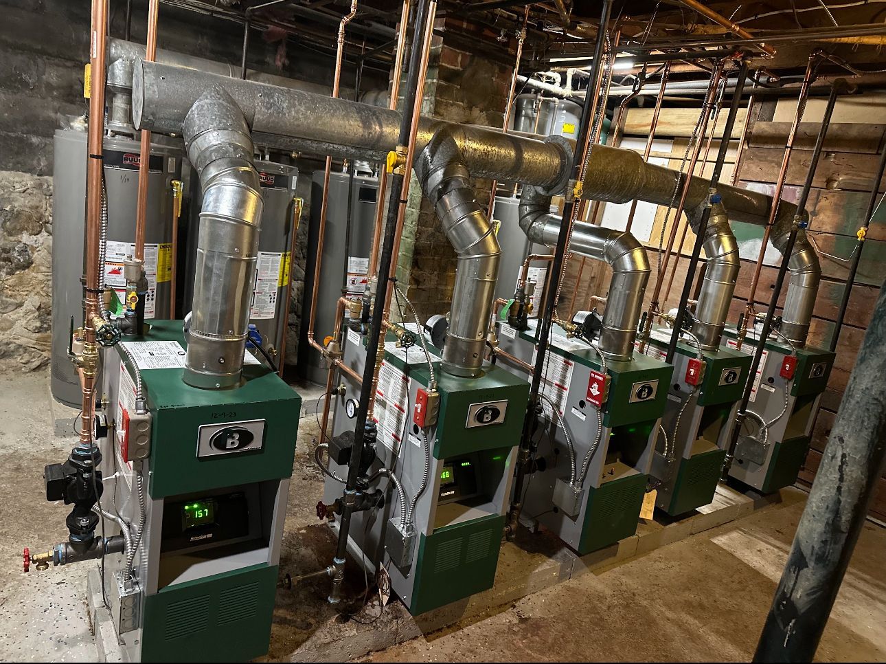Row of green and grey heating boilers with metal pipes, in a basement.