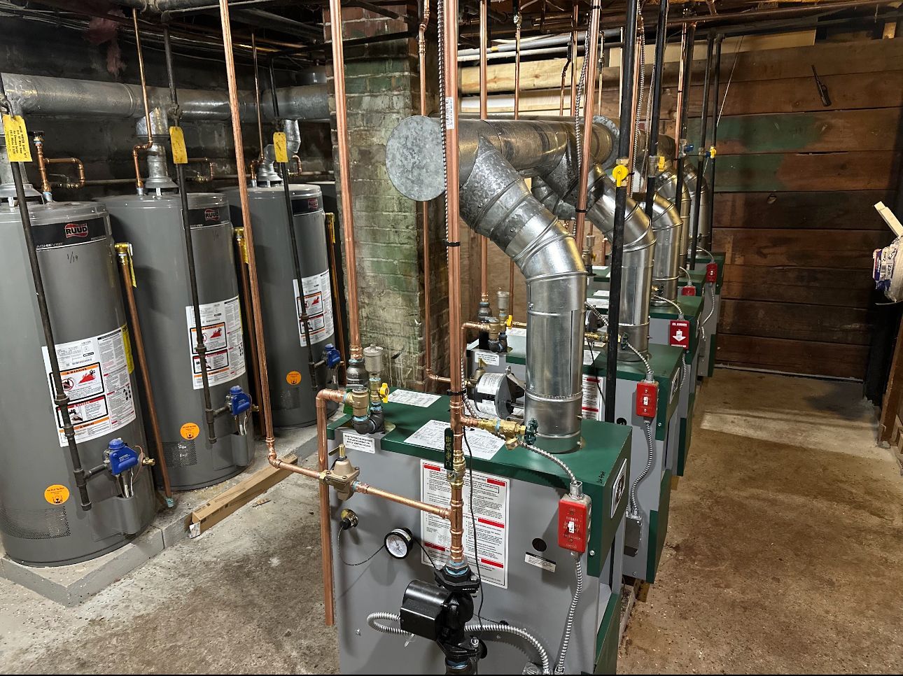 Row of grey water heaters and green boilers in a utility room, connected by copper pipes.