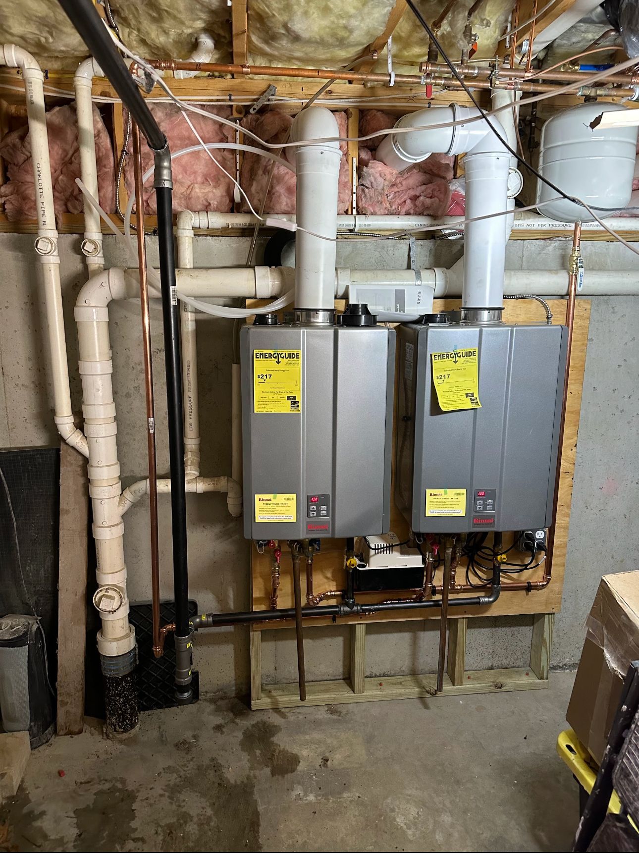 Two silver tankless water heaters mounted on a wooden wall in a basement. Copper pipes, insulation, and ductwork visible.