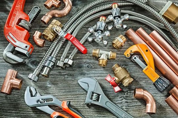 Plumbing tools and copper pipes scattered on a wooden surface, including wrenches, a pipe cutter, and flexible hoses.