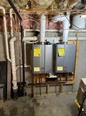 Two silver tankless water heaters mounted on wooden paneling in a basement. Copper and PVC pipes are visible.