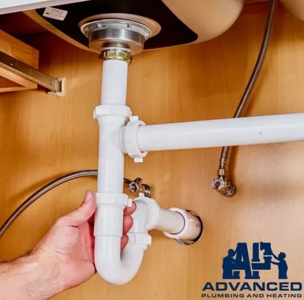 Hand adjusting a white plastic P-trap pipe under a wooden sink; Advanced Plumbing and Heating logo.