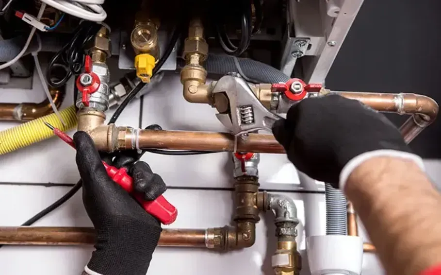 Person in black gloves using a wrench and screwdriver on copper pipes near a boiler.