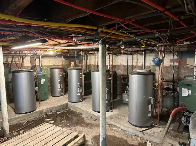 Basement with multiple large silver water heaters and exposed pipes. Wooden beams and concrete floor visible.