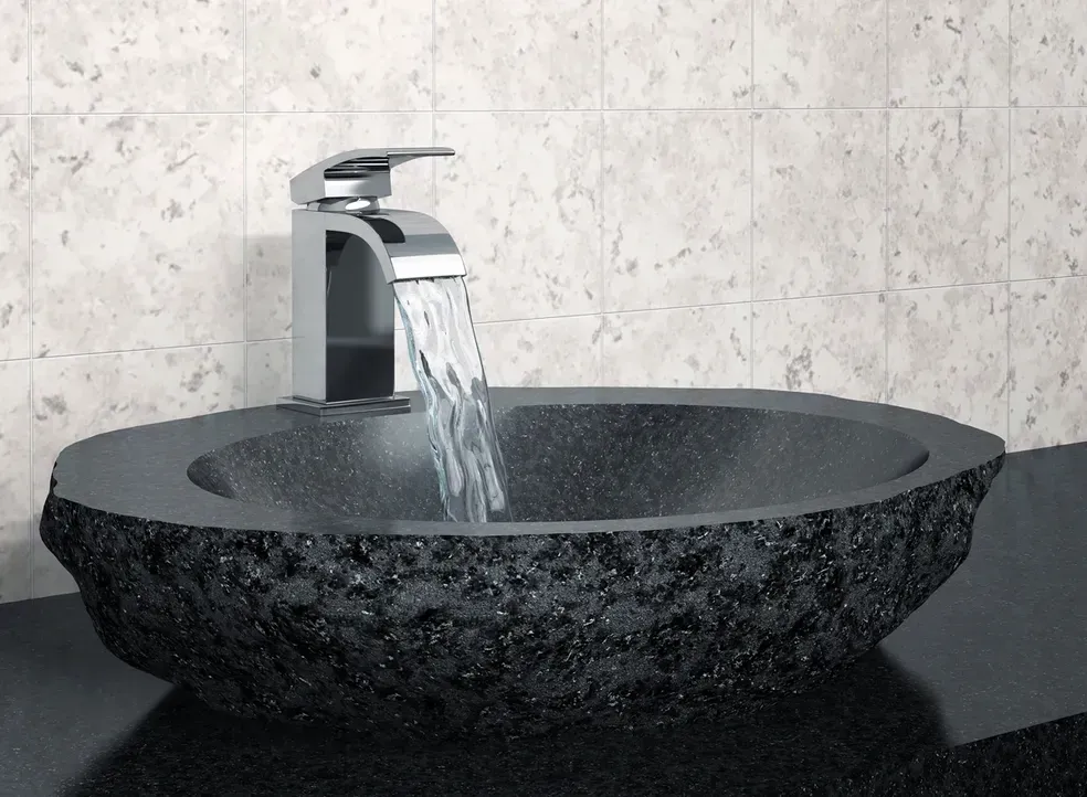 A chrome faucet pours water into a textured black stone vessel sink, set against a tiled wall.