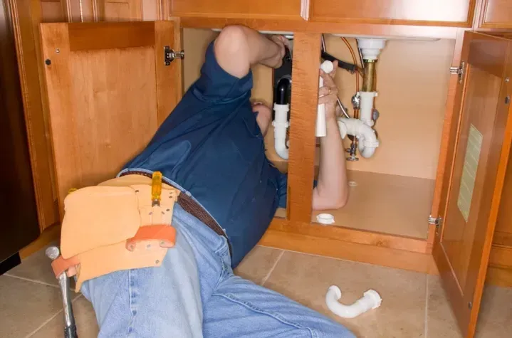 Plumber in blue shirt and jeans working under a kitchen sink, with tools and pipes.