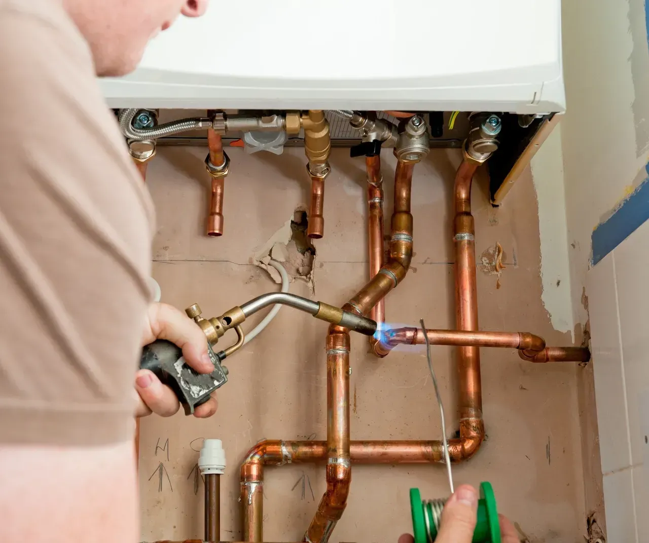 Plumber using a torch to solder copper pipes near a white boiler in a wall.