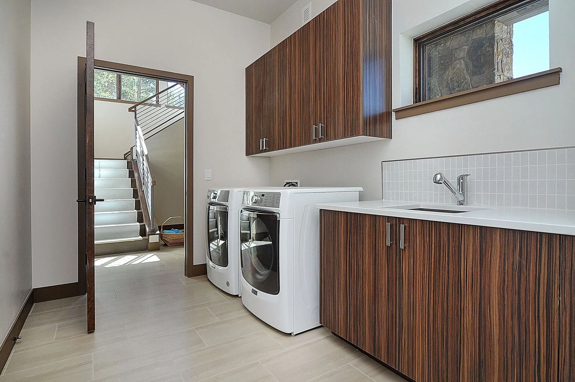 Laundry room with white washer and dryer, wood cabinets, and tiled floor near a staircase.