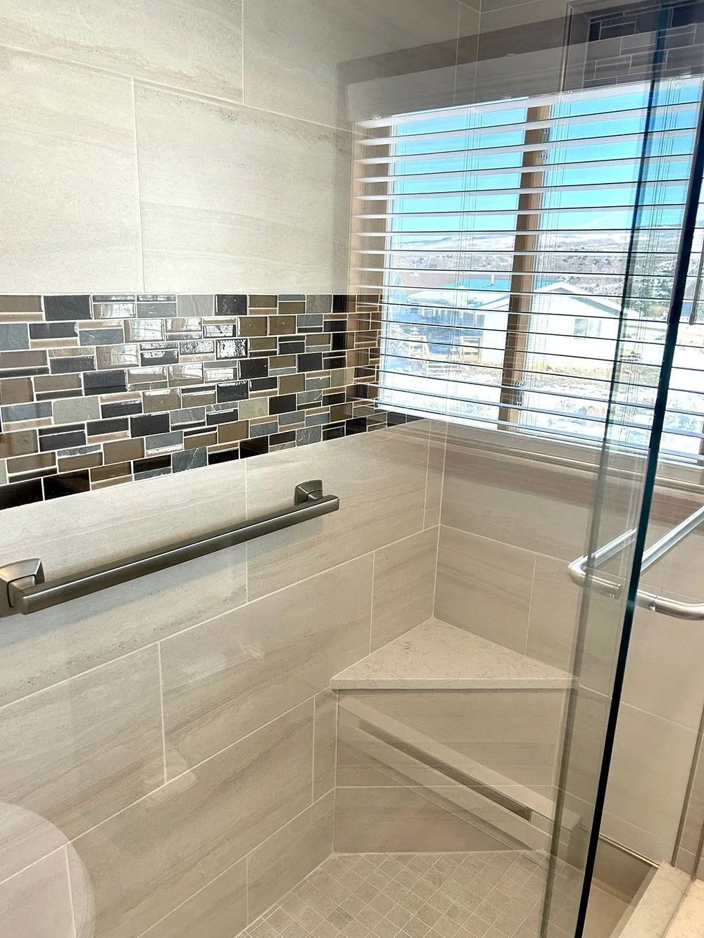 Small tiled bathroom shower with bench, grab bar, mosaic accent strip, and window blinds.
