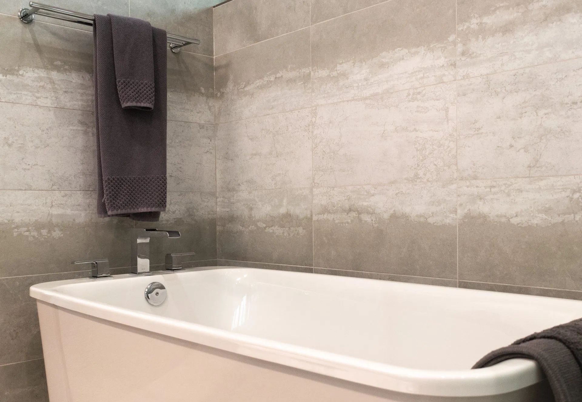 Modern bathroom with a white bathtub, gray tile walls, and dark towels hanging nearby