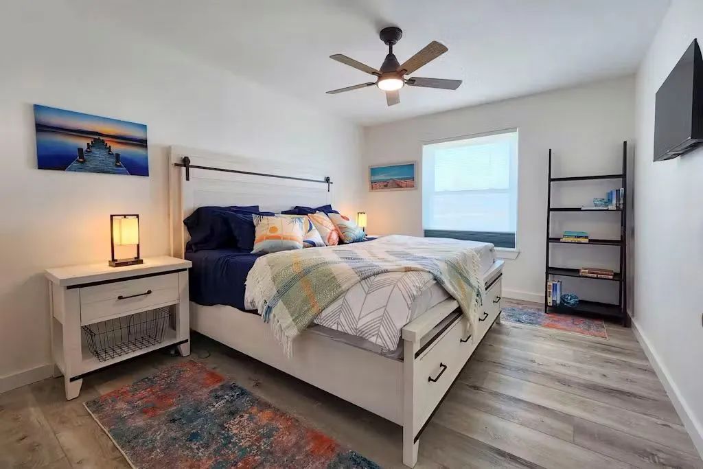 Bright bedroom with a bed, nightstand lamp, window, wall art, ceiling fan, and shelving unit.