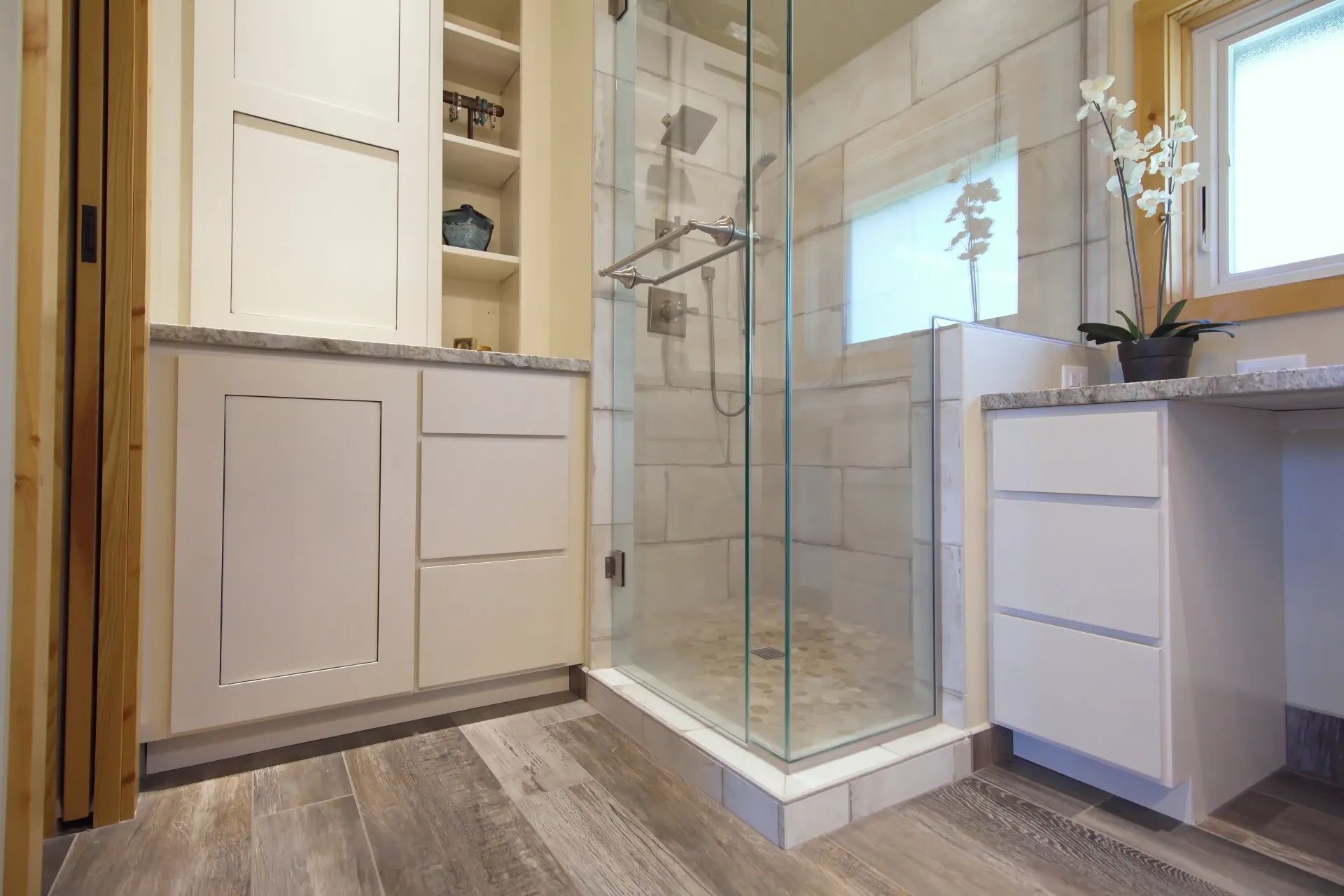Modern bathroom with glass shower, white vanity cabinets, and gray wood-look flooring.