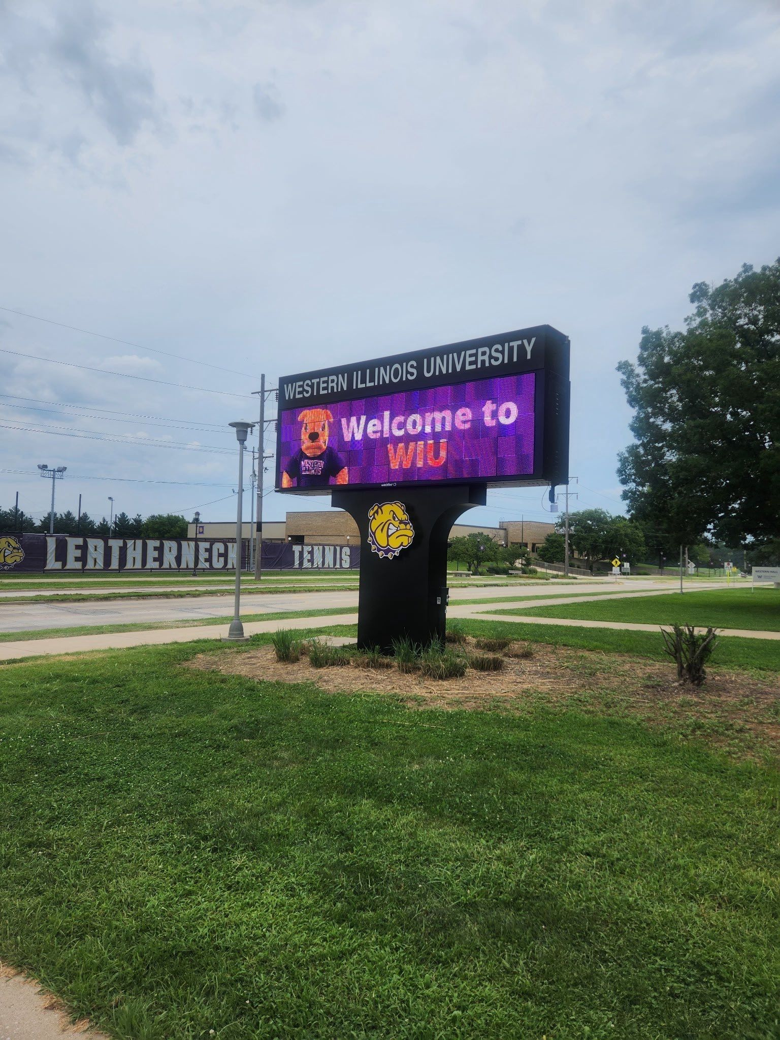 Texas A&M University welcome sign on a grassy roadside under a cloudy sky, with the Aggie logo and digital display.