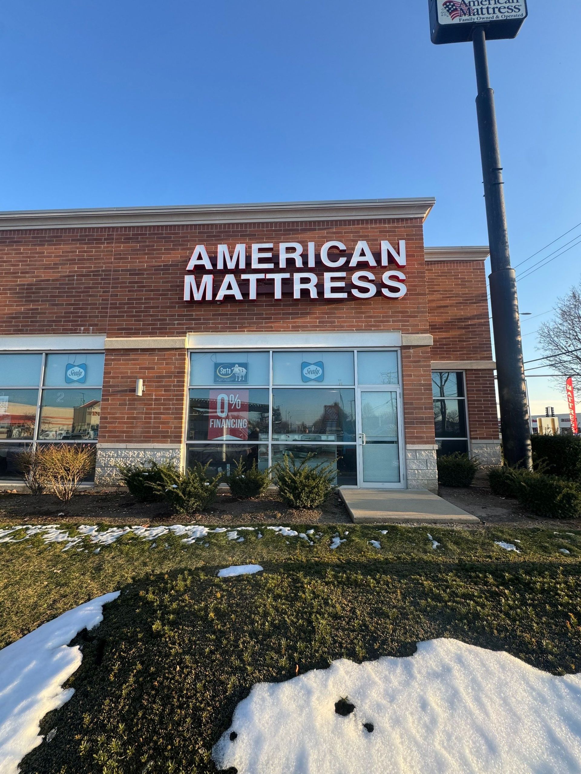 American Mattress storefront with brick facade, glass entrance, and snow-covered landscaping under a blue sky
