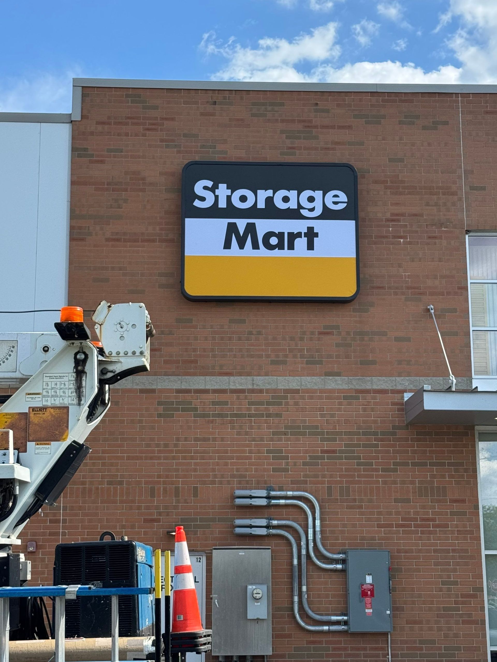 Storage Mart sign on a brick building with utility pipes and a lift basket in the foreground