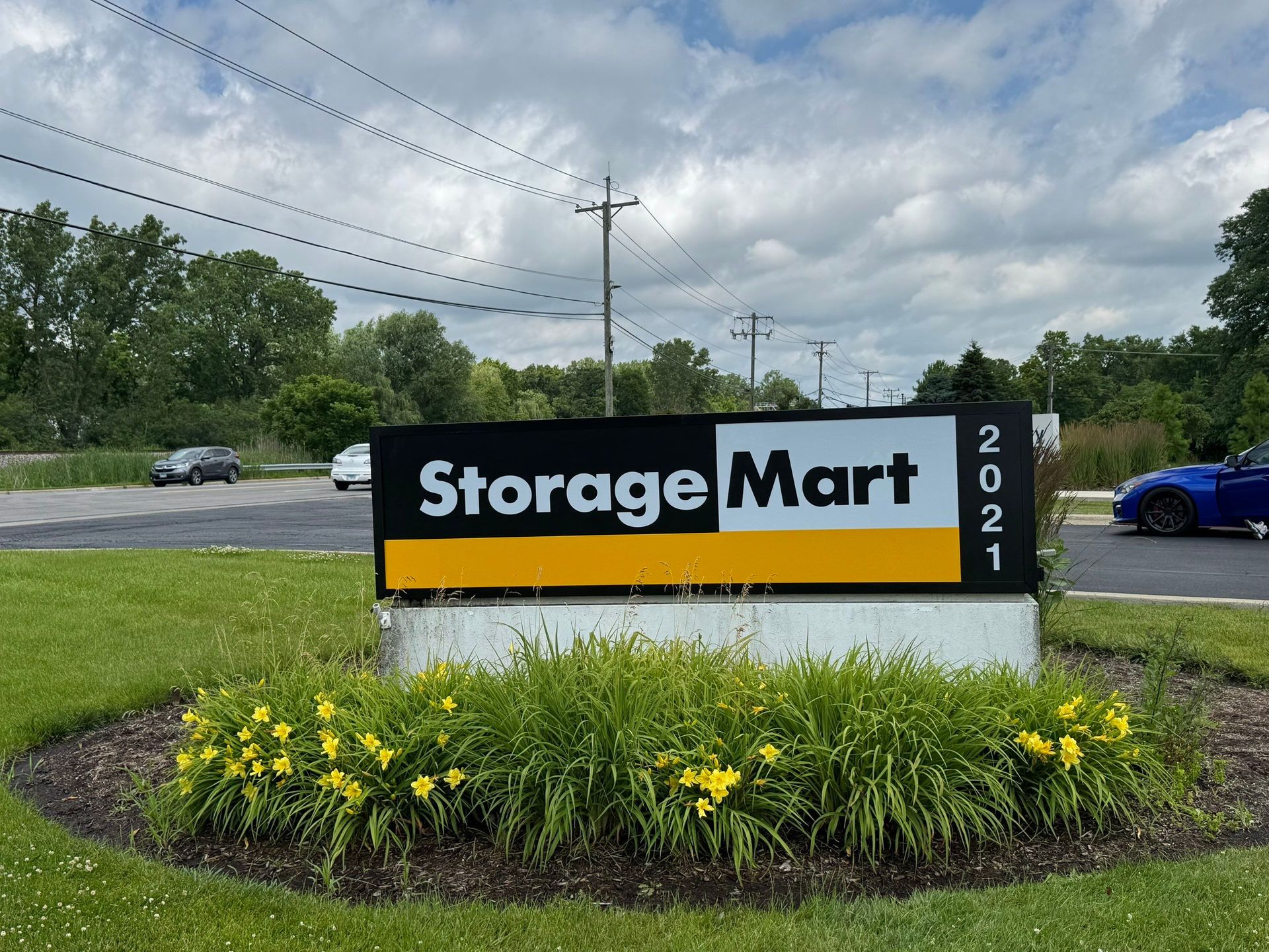 StorageMart roadside sign with yellow flowers, black and orange lettering, and cloudy sky behind it
