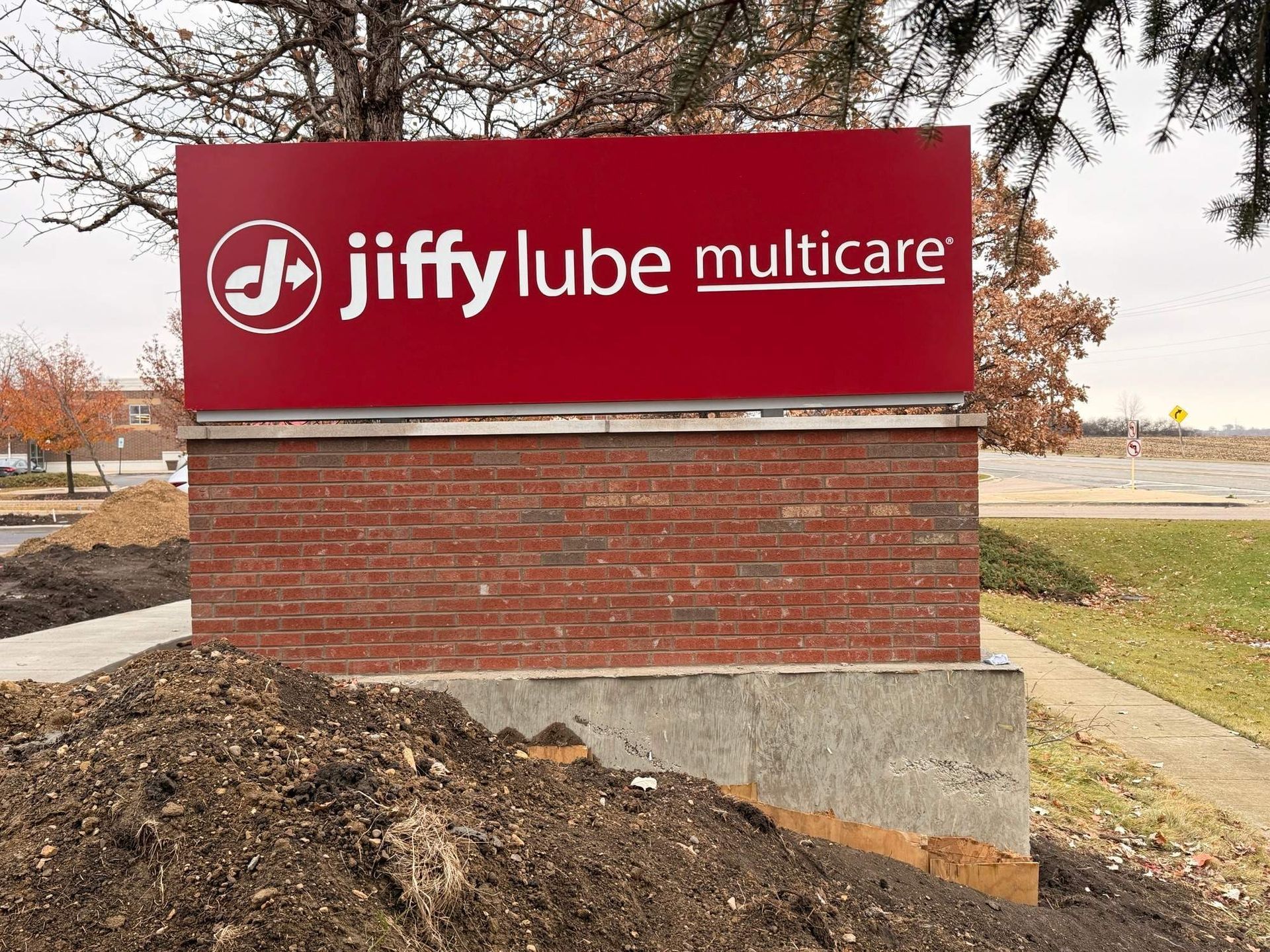 Jiffy Lube Multicare sign on a brick base beside a roadside, with dirt and trees around it