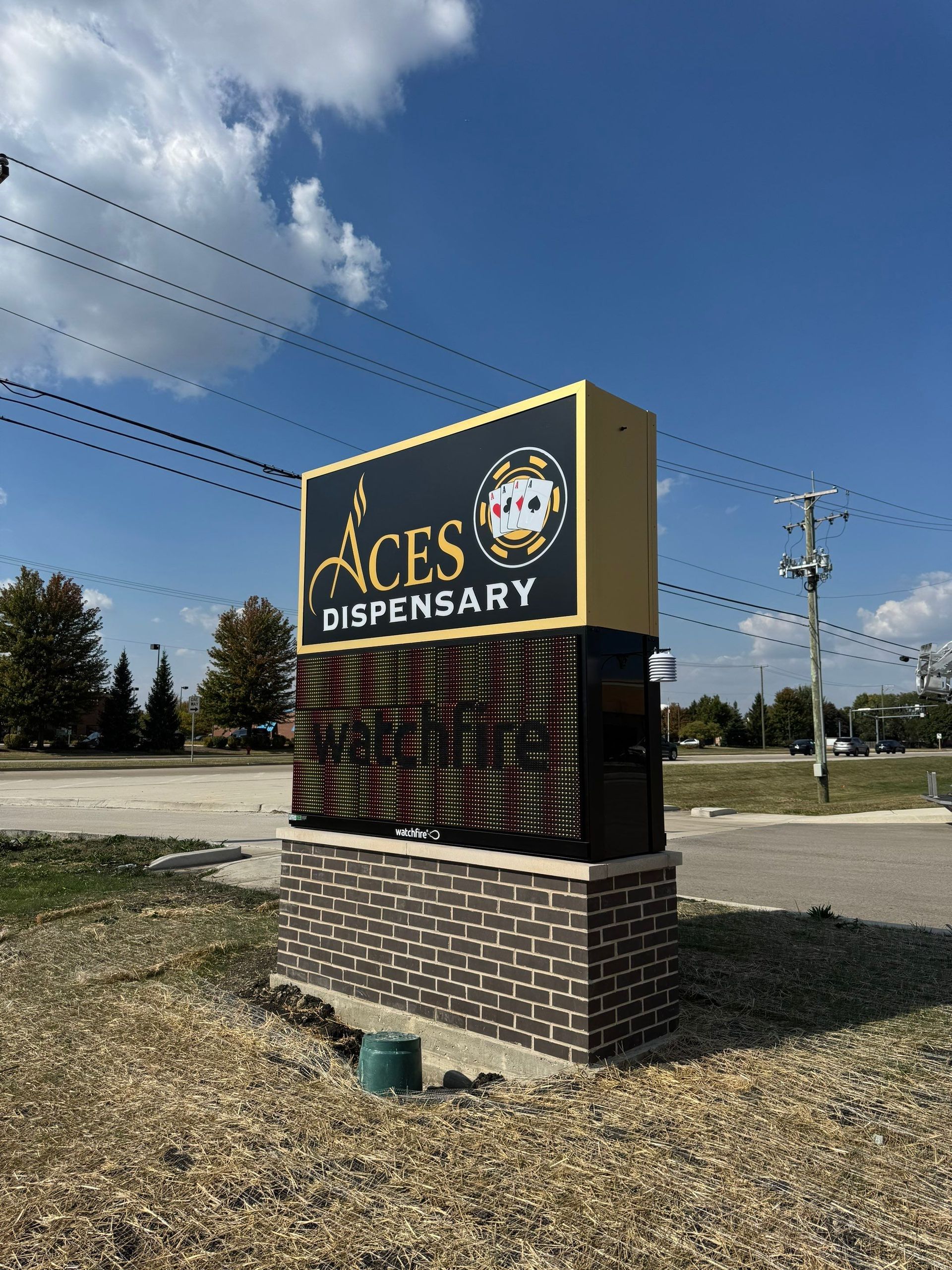 Aces Dispensary roadside sign on a brick base under a blue sky with power lines.