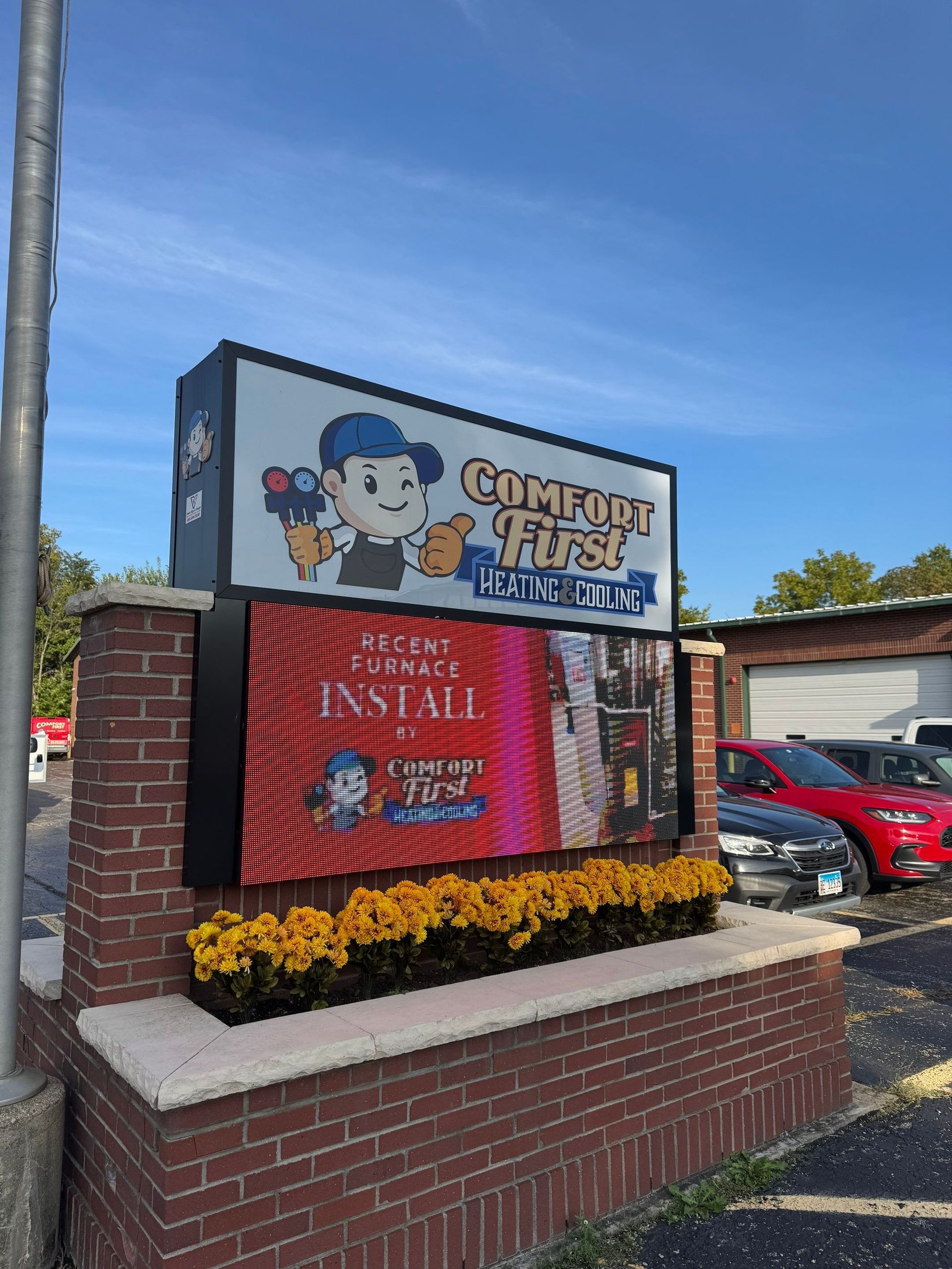 Crumbl Cookies sign with mascot, red digital menu board, and flower bed outside a brick storefront.