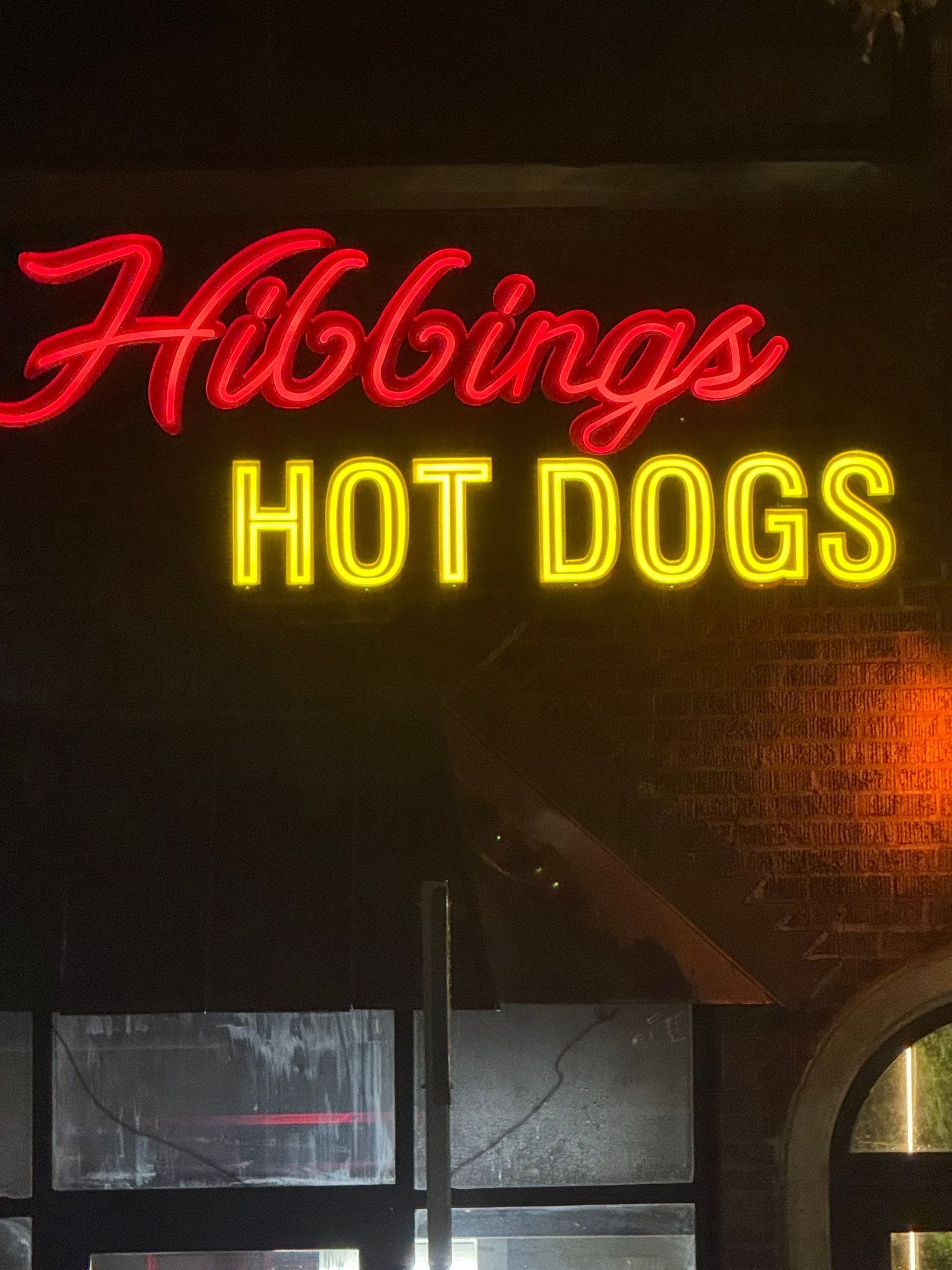 Neon sign for “Hibbings HOT DOGS” glowing red and yellow on a dark storefront at night