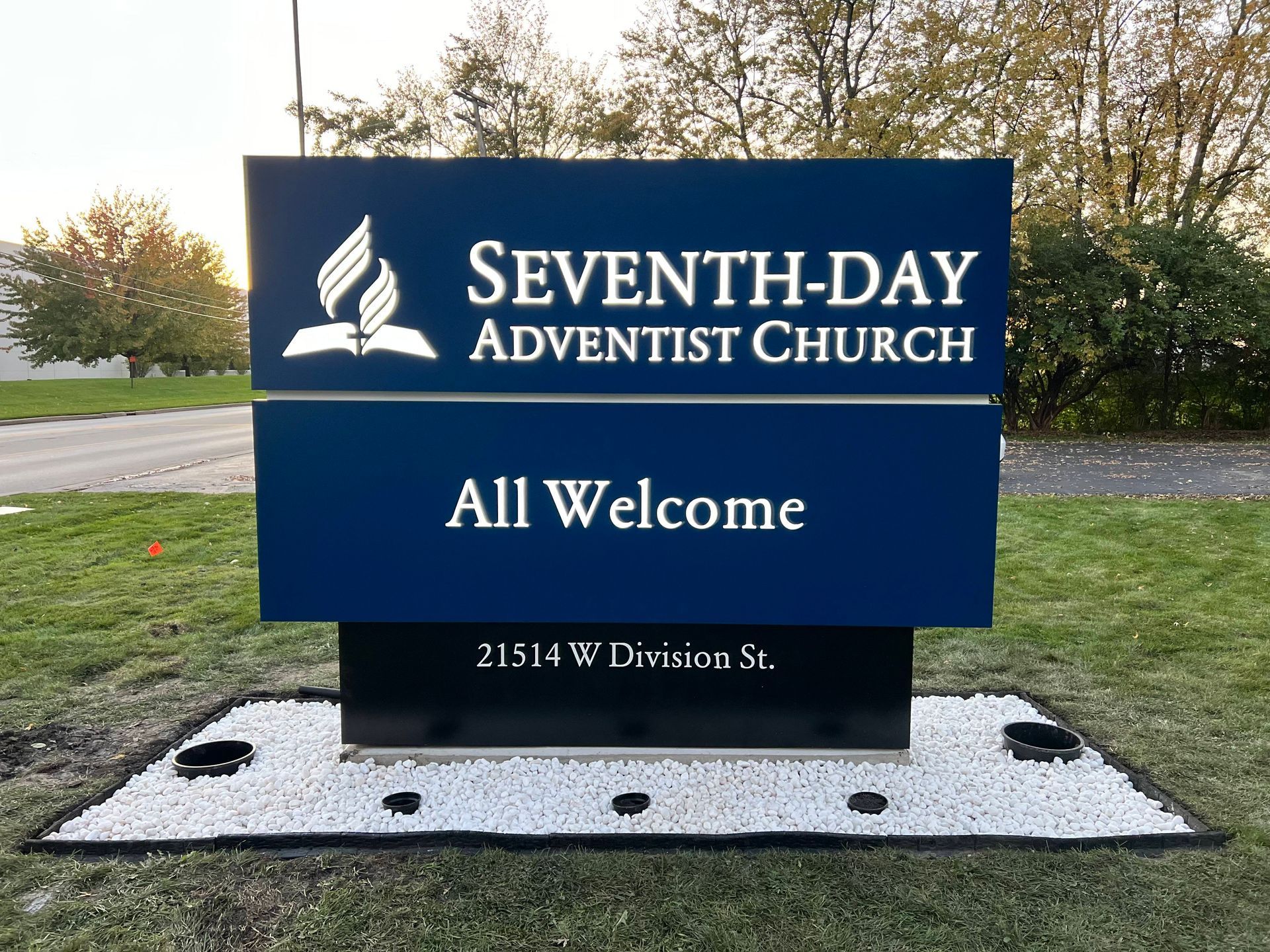 Seventh-day Adventist Church sign on a lawn, blue with “All Welcome” and address 21514 W Division St.