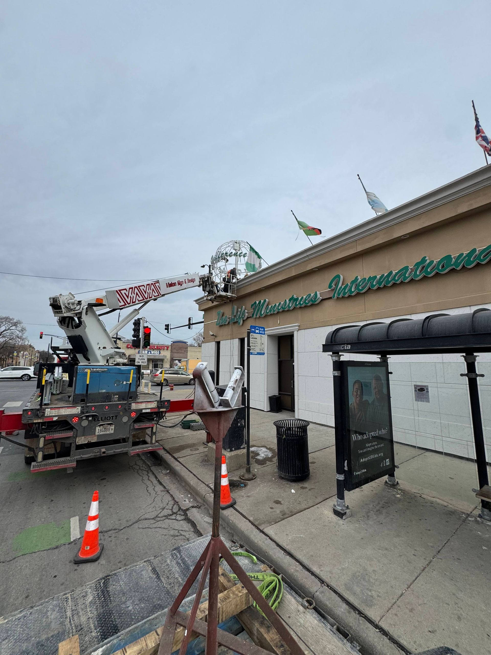 Bucket truck with extended boom near a building entrance on a cloudy street, with traffic cones and a ladder.