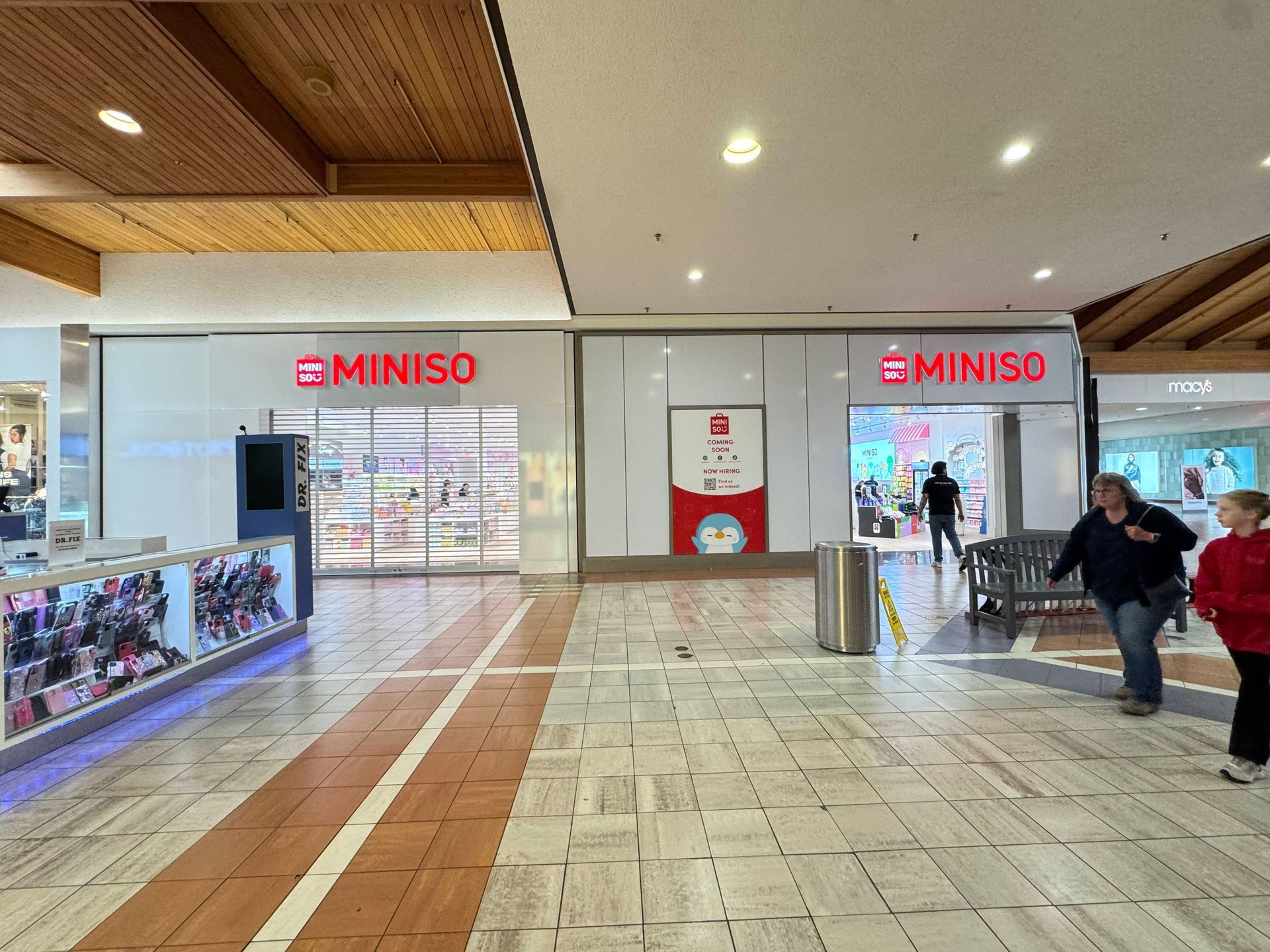 MINISO store entrance in a mall, with bright lights, display cases, and two shoppers walking by.