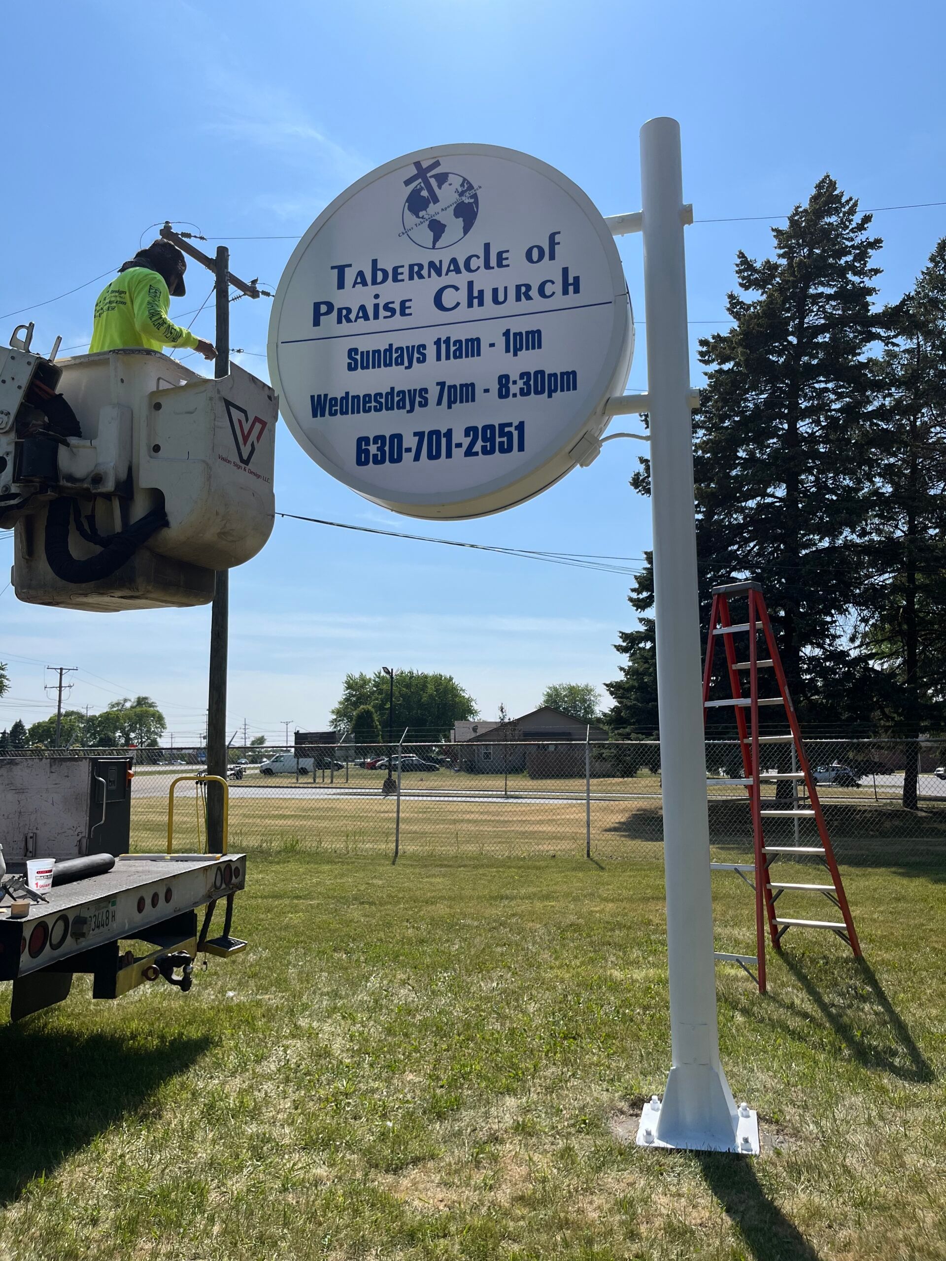 White sign for Tabernacle of Praise Church with service times beside a roadside and ladder in a grassy field